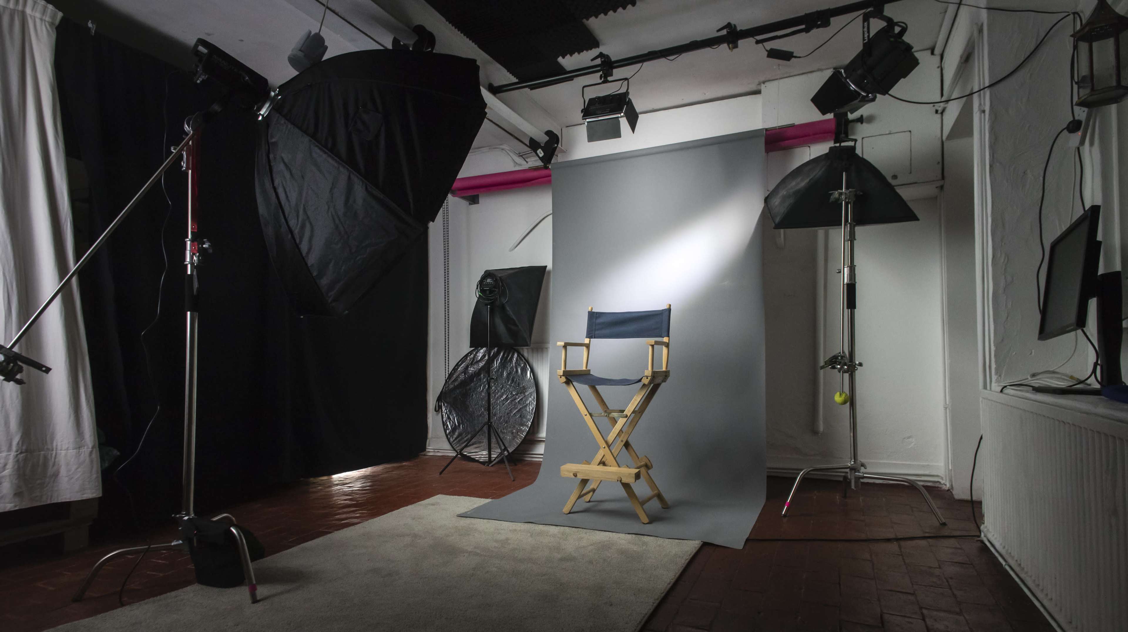 A photography studio setup with a director's chair positioned in front of a gray backdrop, surrounded by various lighting equipment and a carpeted area.