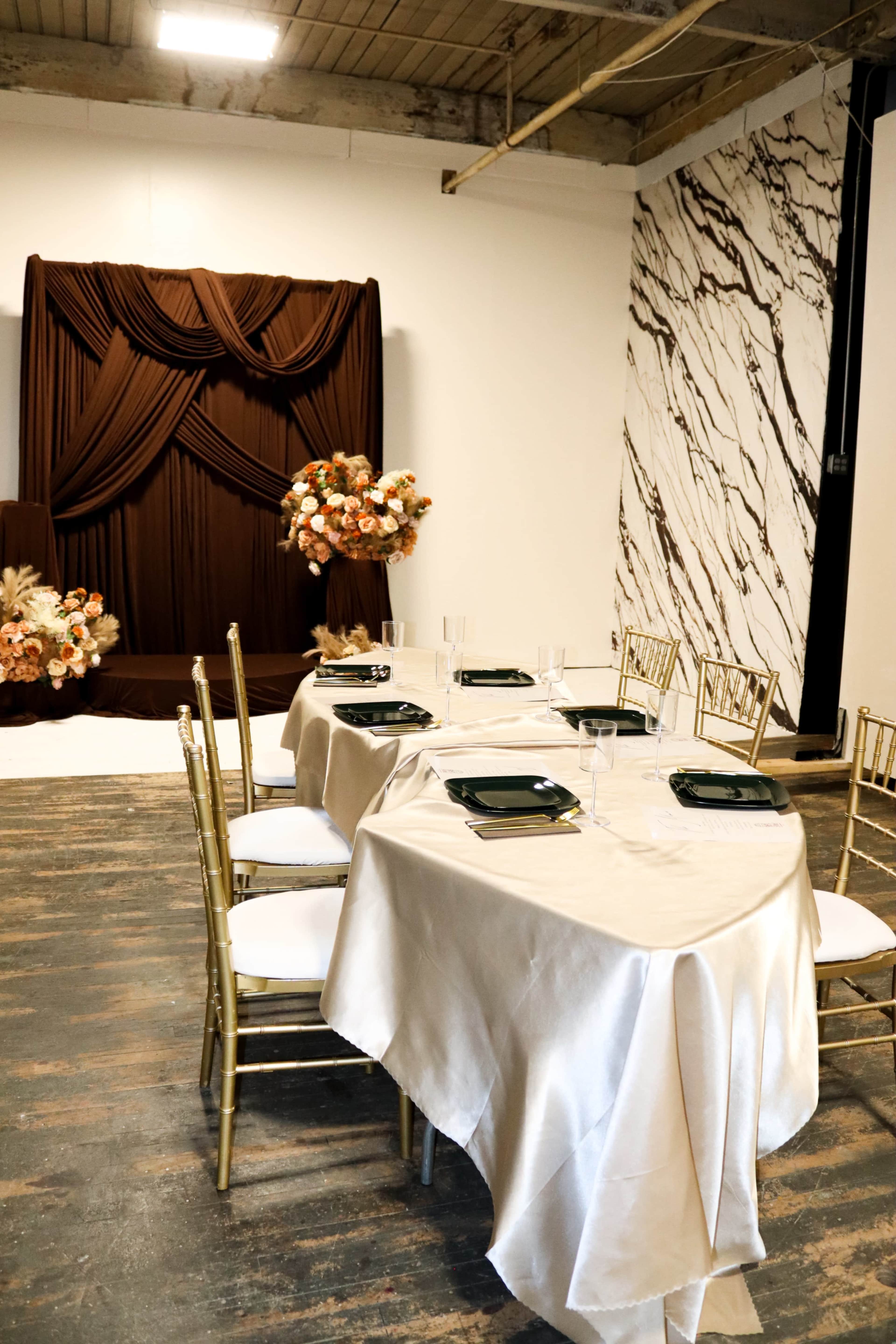 A table is set with elegant tableware in front of a backdrop featuring draped fabric and floral arrangements, all situated in a modern space with marble-patterned walls.