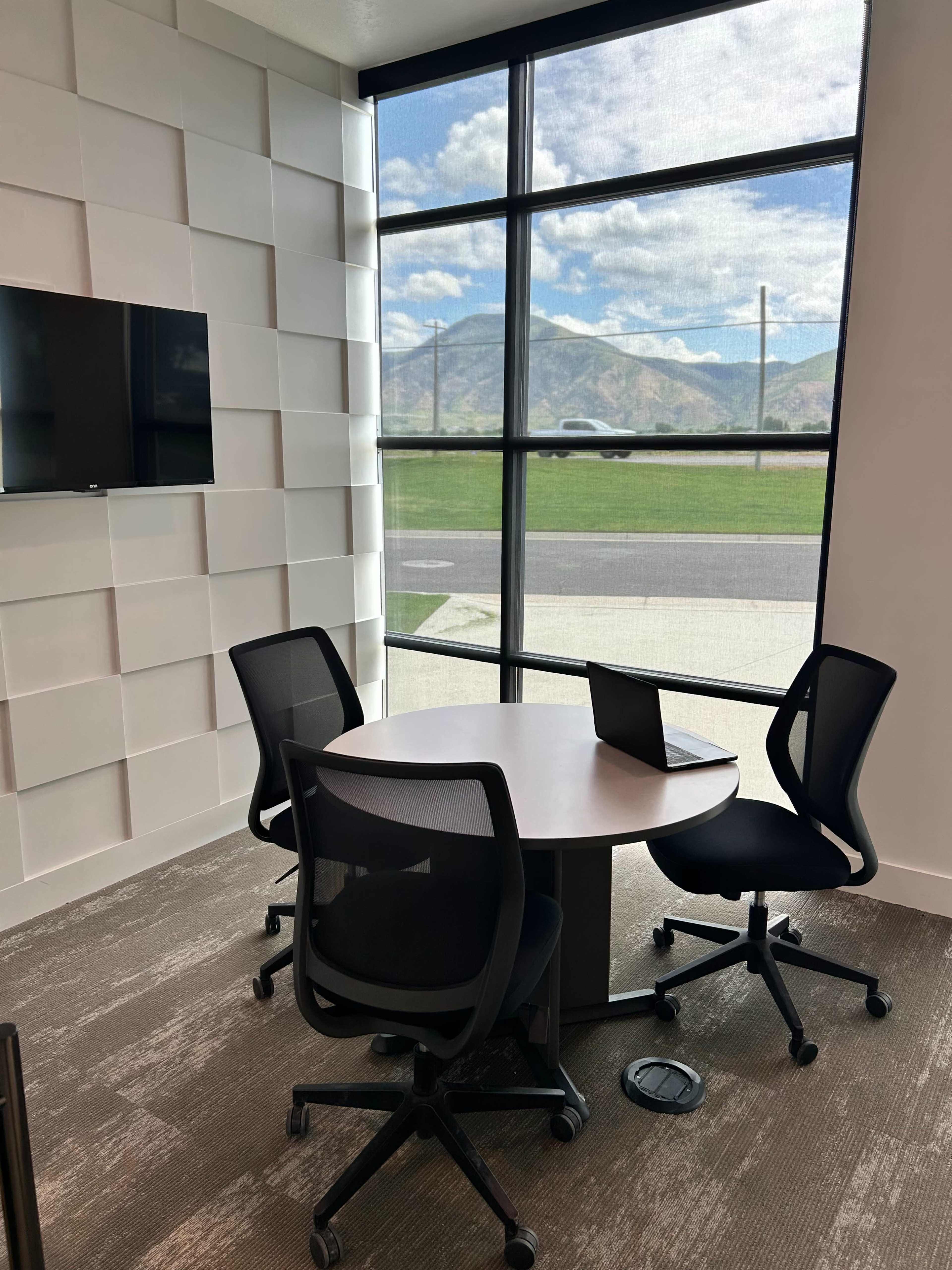 A conference room features a round table with three chairs, a wall-mounted TV, and large windows showcasing a view of mountains and greenery.