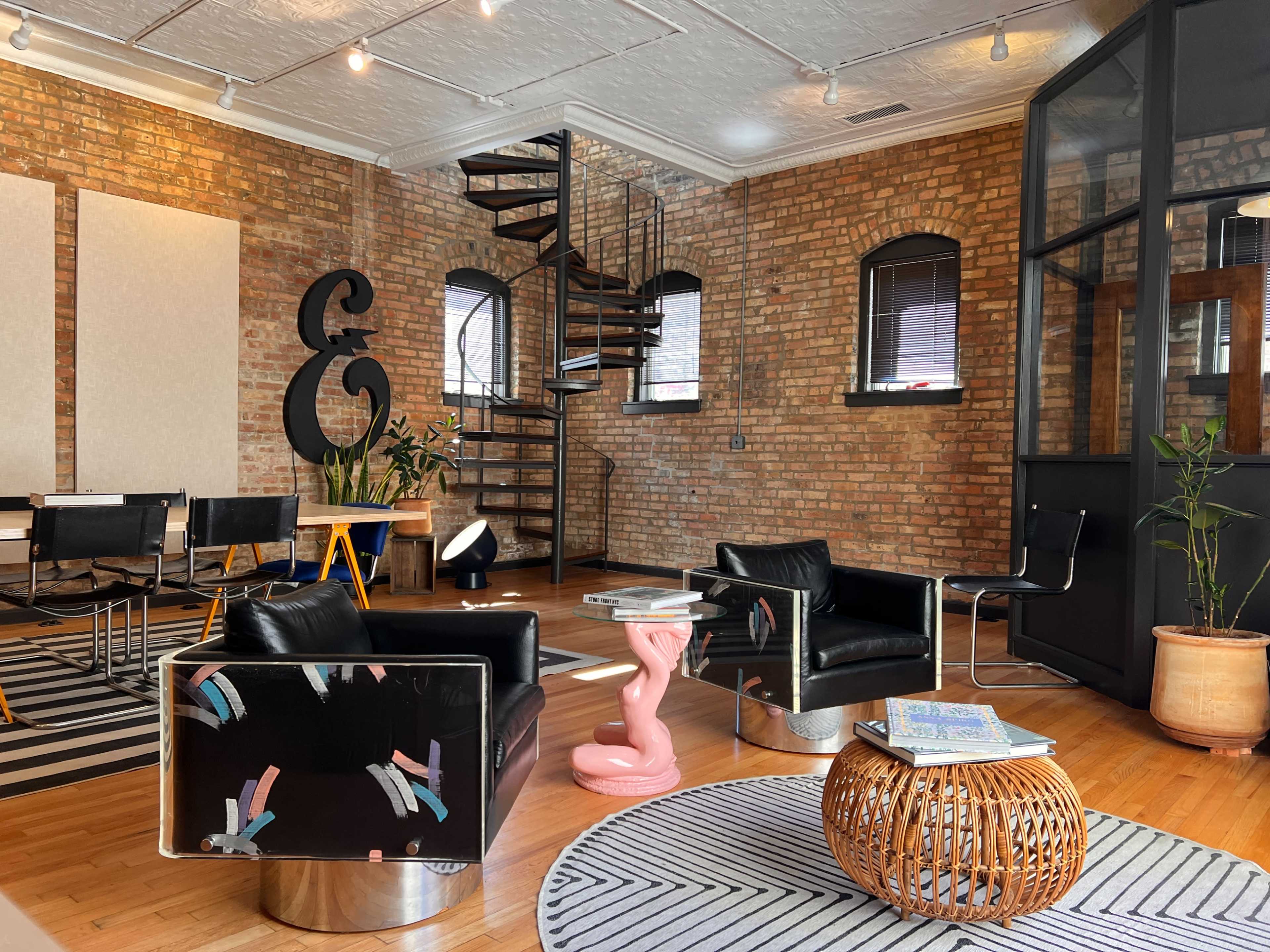 The image shows a stylish interior with exposed brick walls, modern furniture, and a spiral staircase.