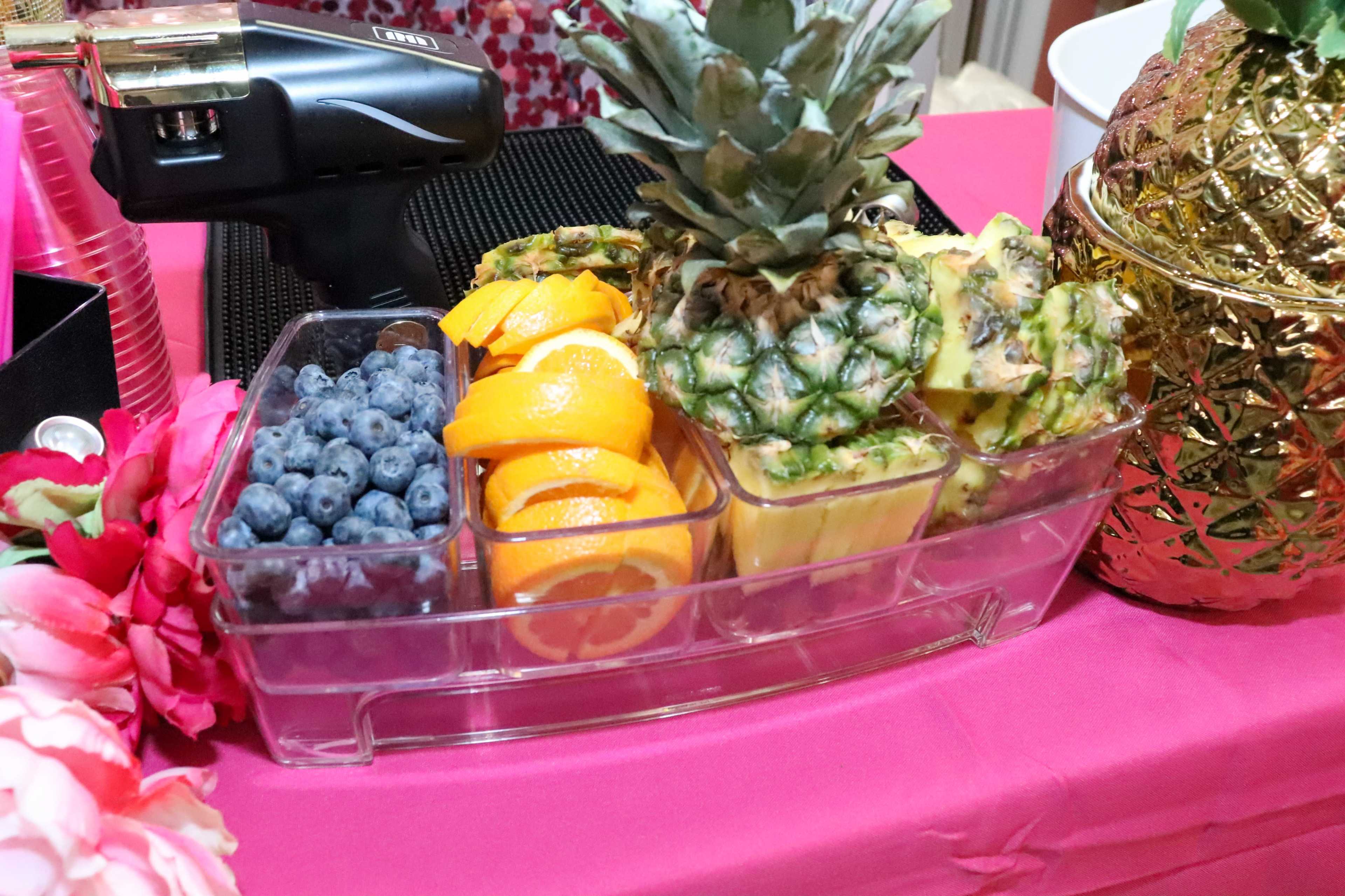 A clear plastic container holds sliced oranges, blueberries, and pineapple arranged on a pink table.