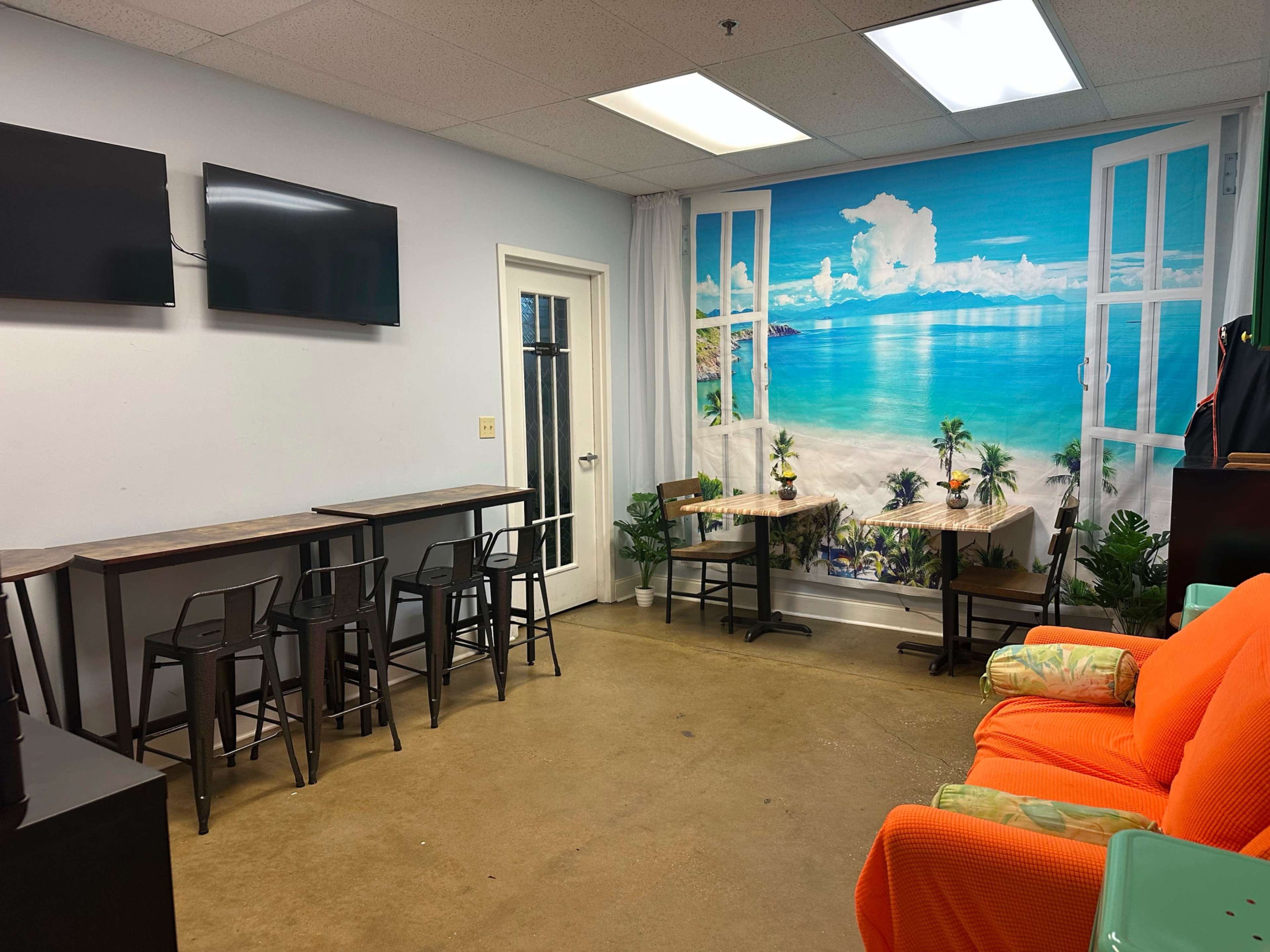 The image shows a small room with two wall-mounted televisions, several tall tables and chairs, and a vibrant mural of a beach scene on one wall.