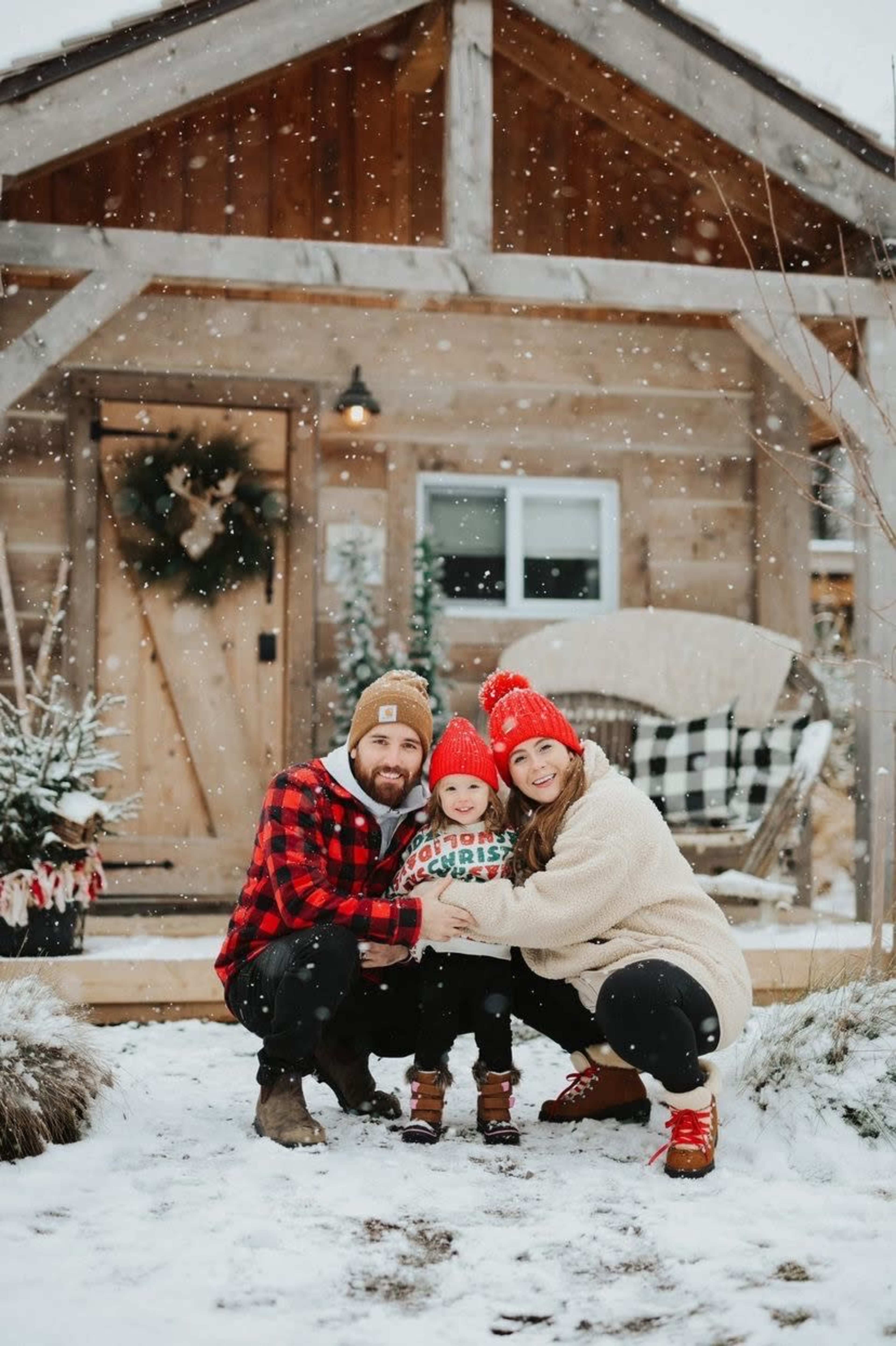 A family of three poses together in the snow in front of a rustic wooden house during a snowfall.