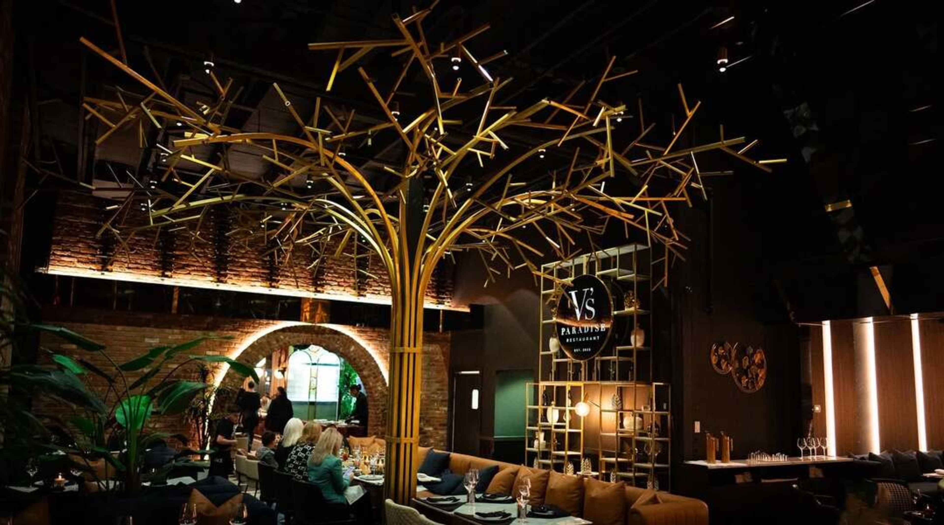 The image shows a restaurant interior featuring a large decorative tree made of bamboo, with tables arranged around it and patrons dining.