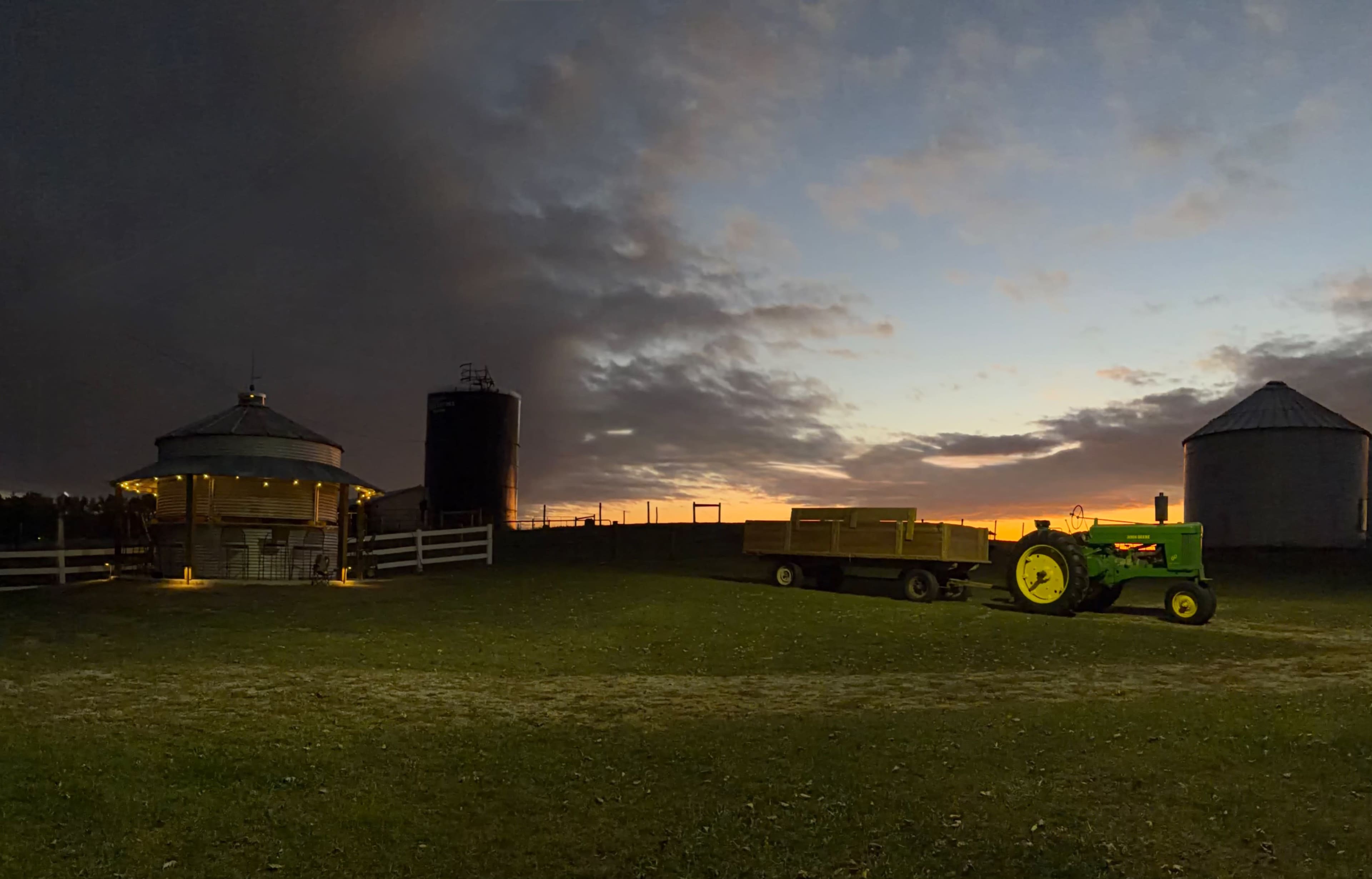 A green tractor and a trailer are positioned on a farm at dusk, with a barn and silos in the background under a cloudy sky.