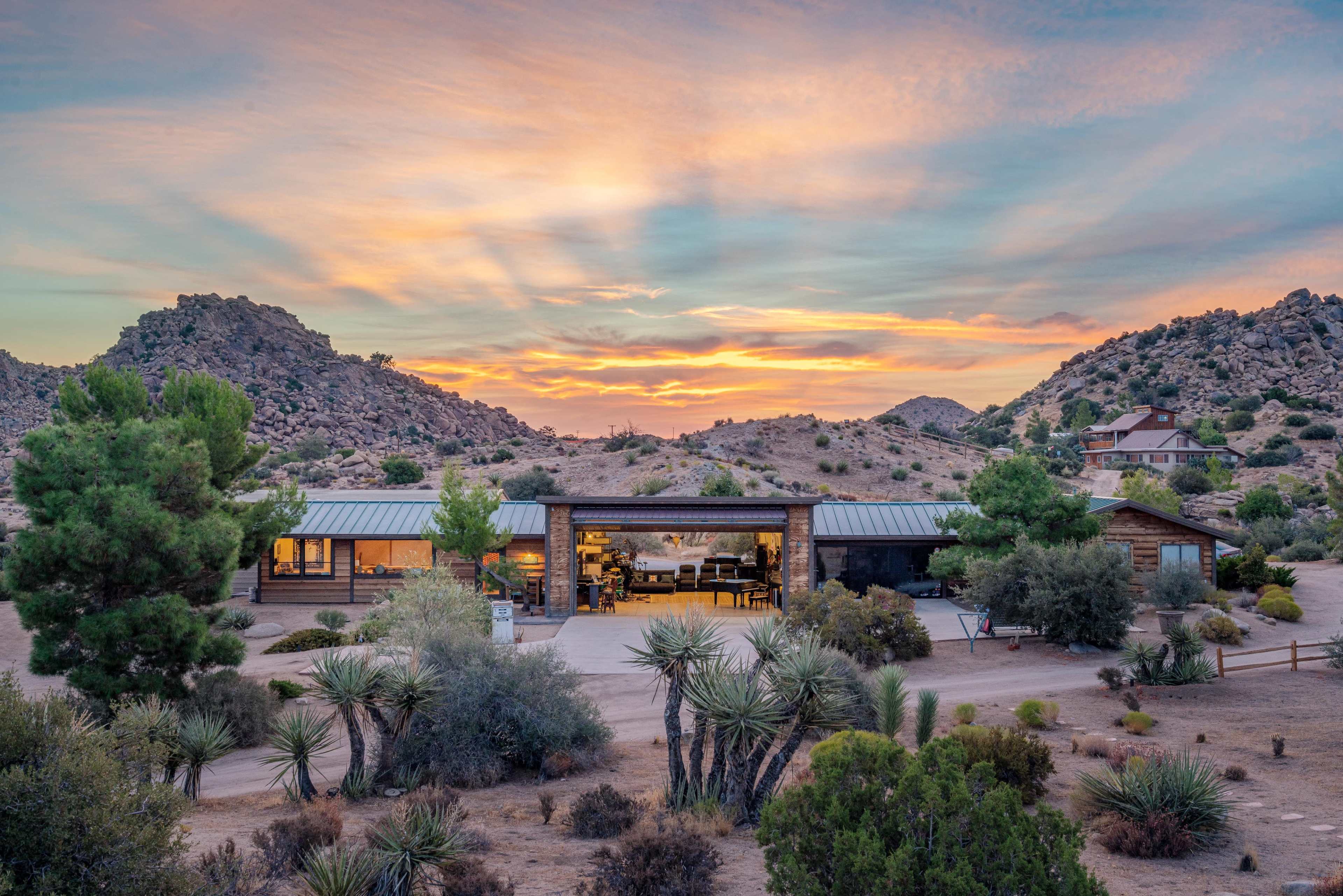 A modern house set against a backdrop of mountains and a colorful sunset, surrounded by desert vegetation.