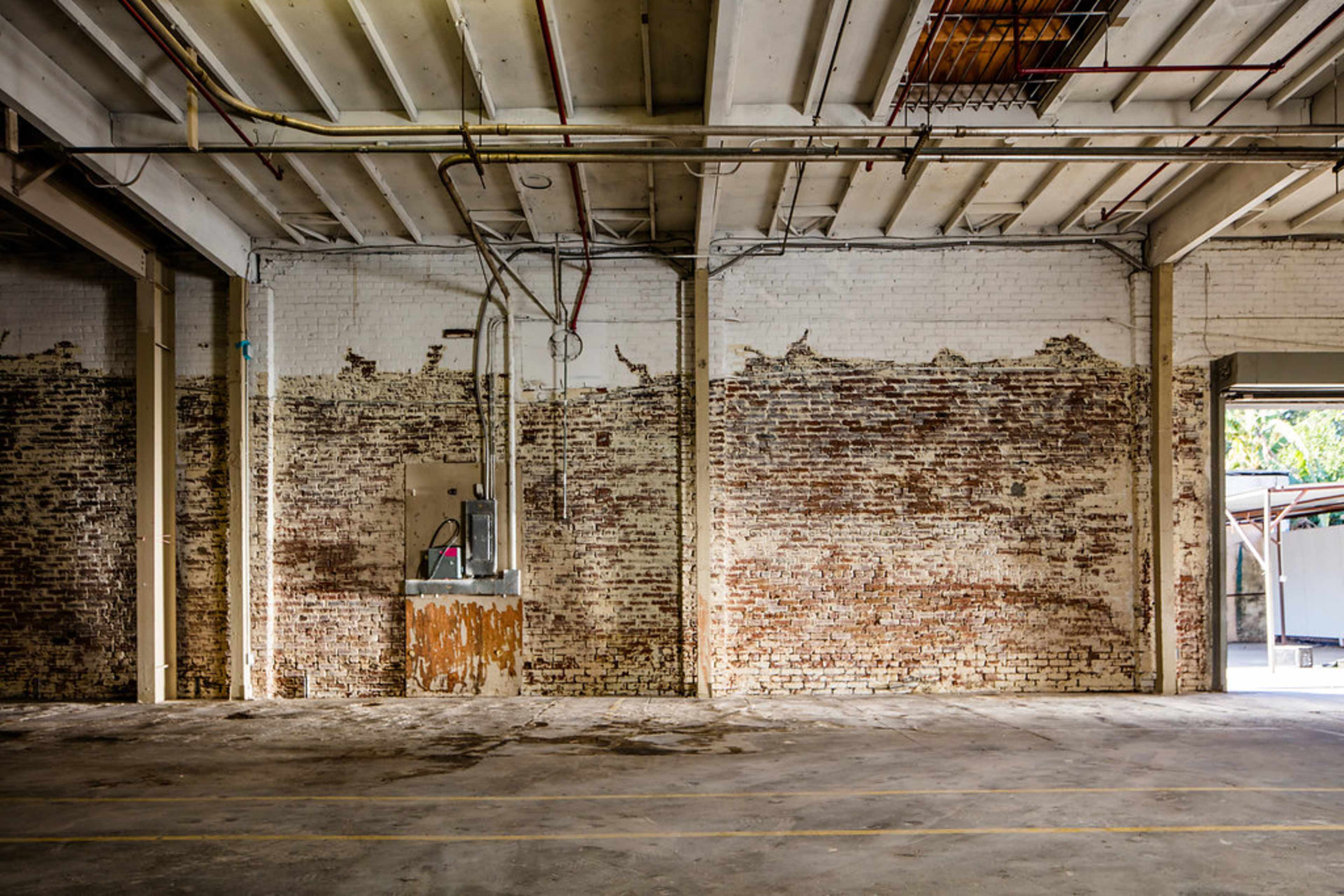 The image shows an empty warehouse interior featuring exposed brick walls and overhead utilities.