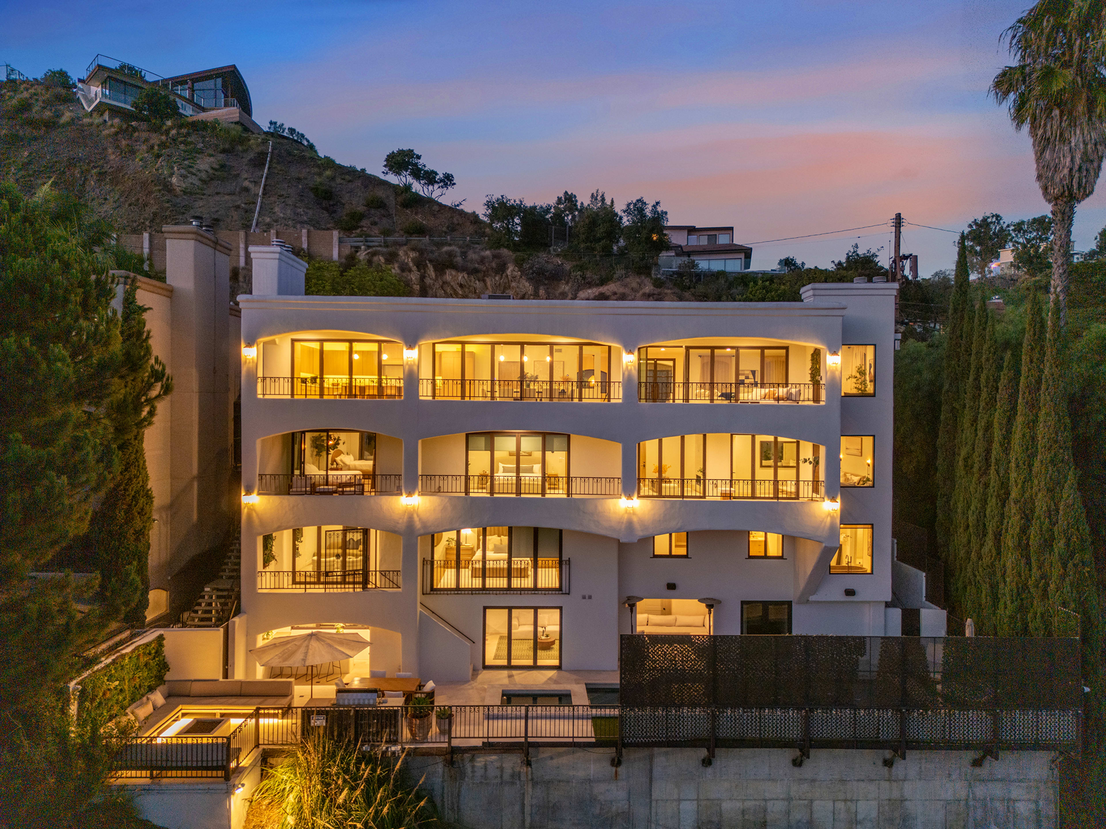 A modern multi-level house with large windows and multiple balconies sits on a hillside, illuminated against a twilight sky.