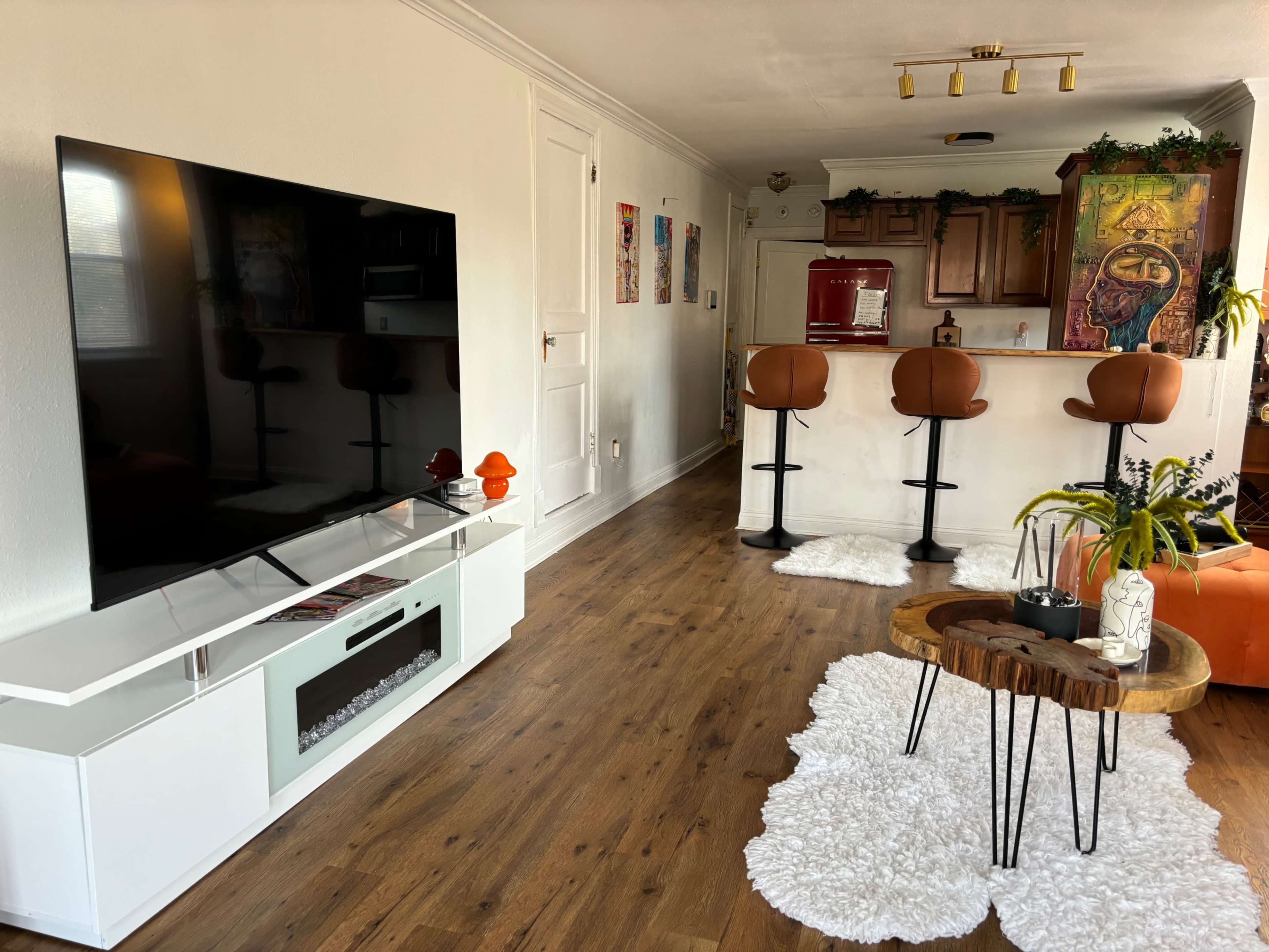 The living room features a large television on a white media unit, with a bar area behind it that includes two brown barstools and a wooden coffee table on a white rug.