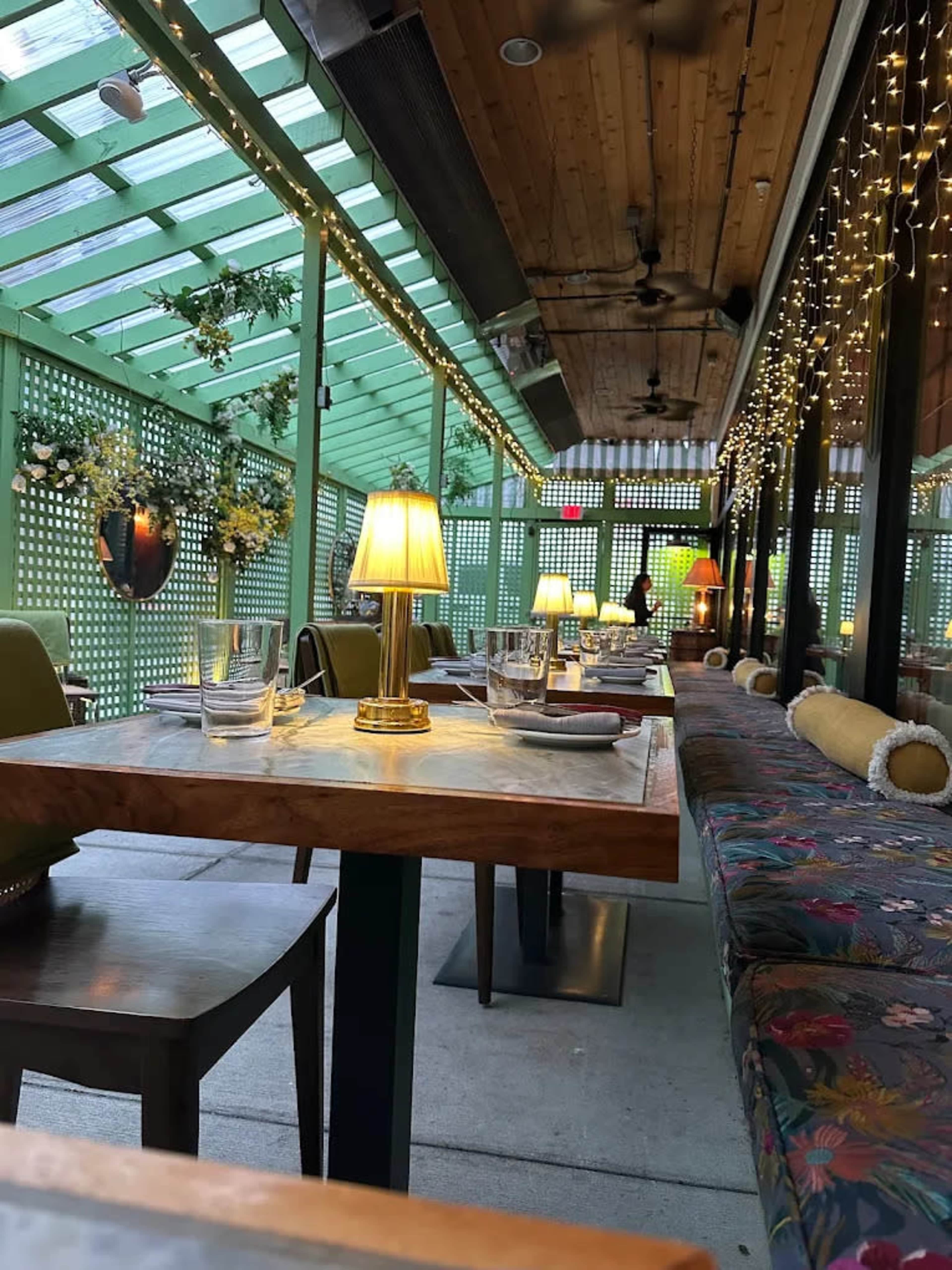 The image shows a modern restaurant with indoor and outdoor seating, featuring tables adorned with lamps and surrounded by greenery and decorative lighting.