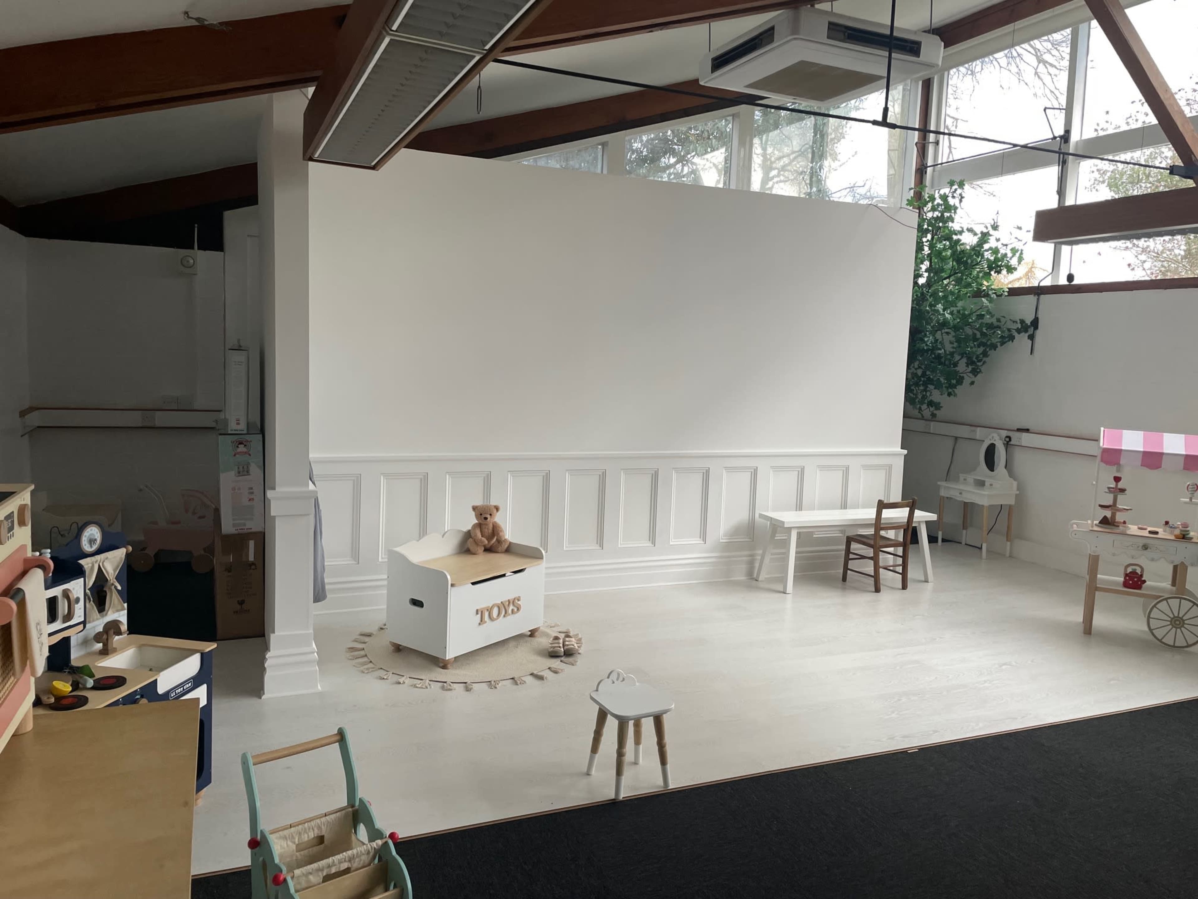 A spacious room with a white wall, wooden beams, and minimal furniture, including a toy box, a small table, and a chair.
