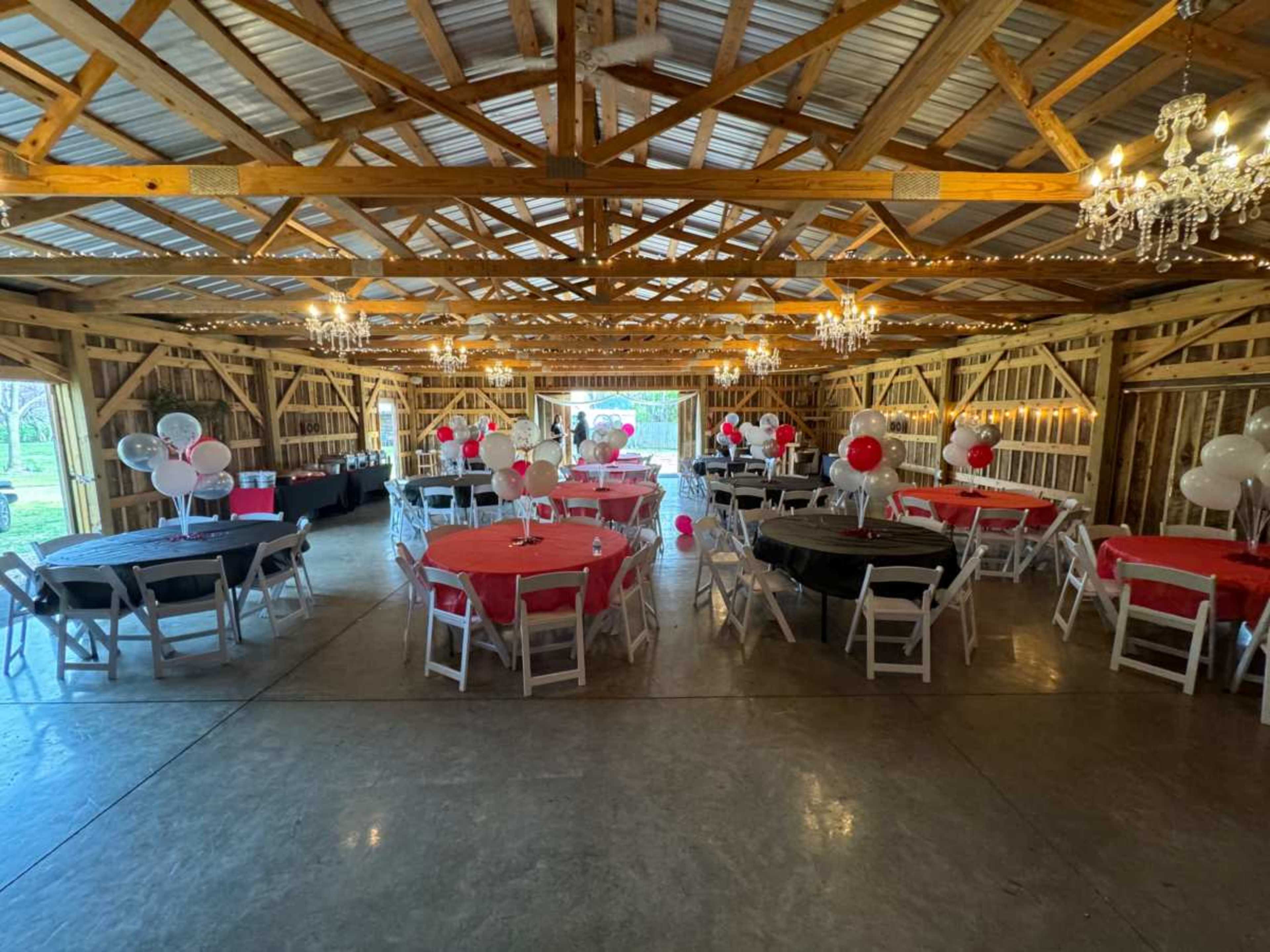 The interior of a barn is decorated for an event, featuring round tables with red and black tablecloths, balloons, and chandeliers hanging from the ceiling.