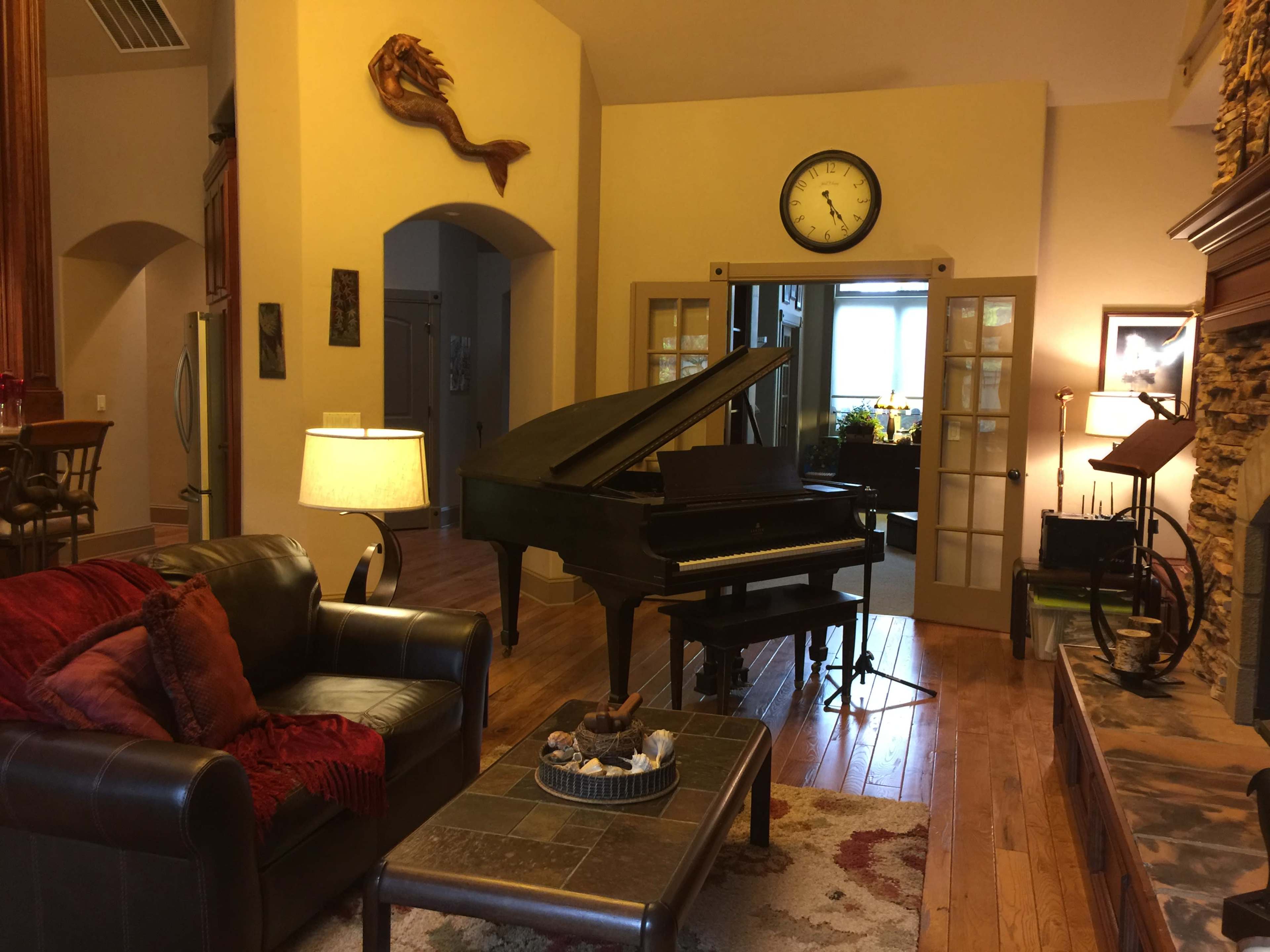 A cozy living space featuring a grand piano, a leather sofa with red throw pillows, and a stone fireplace, complemented by warm lighting and wooden floors.