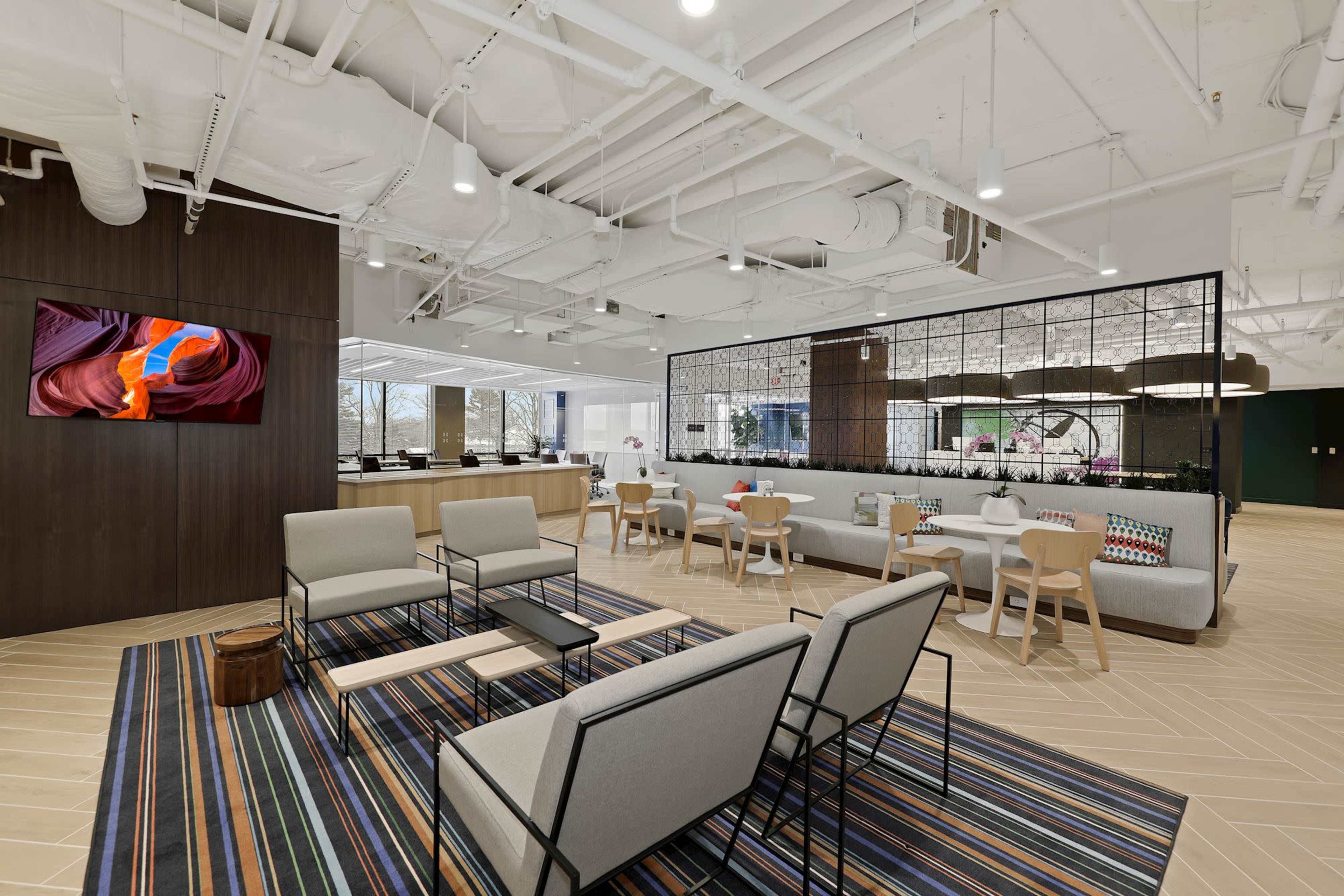 The image shows a modern office lounge area with seating, a large screen displaying colorful artwork, and a glass partition with plants.