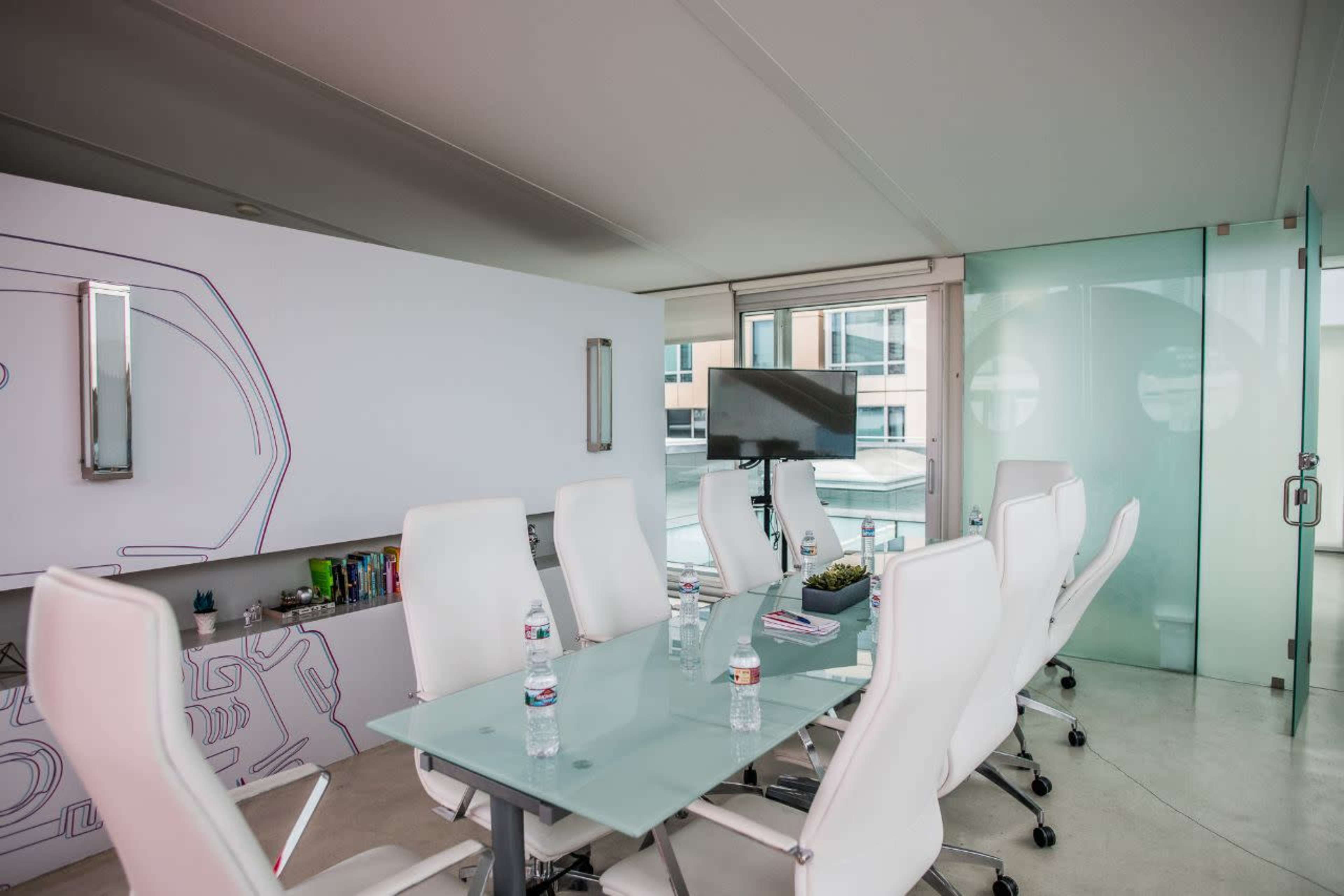 A modern conference room features a glass table surrounded by white chairs, with a wall displaying artwork and a television screen on one side.