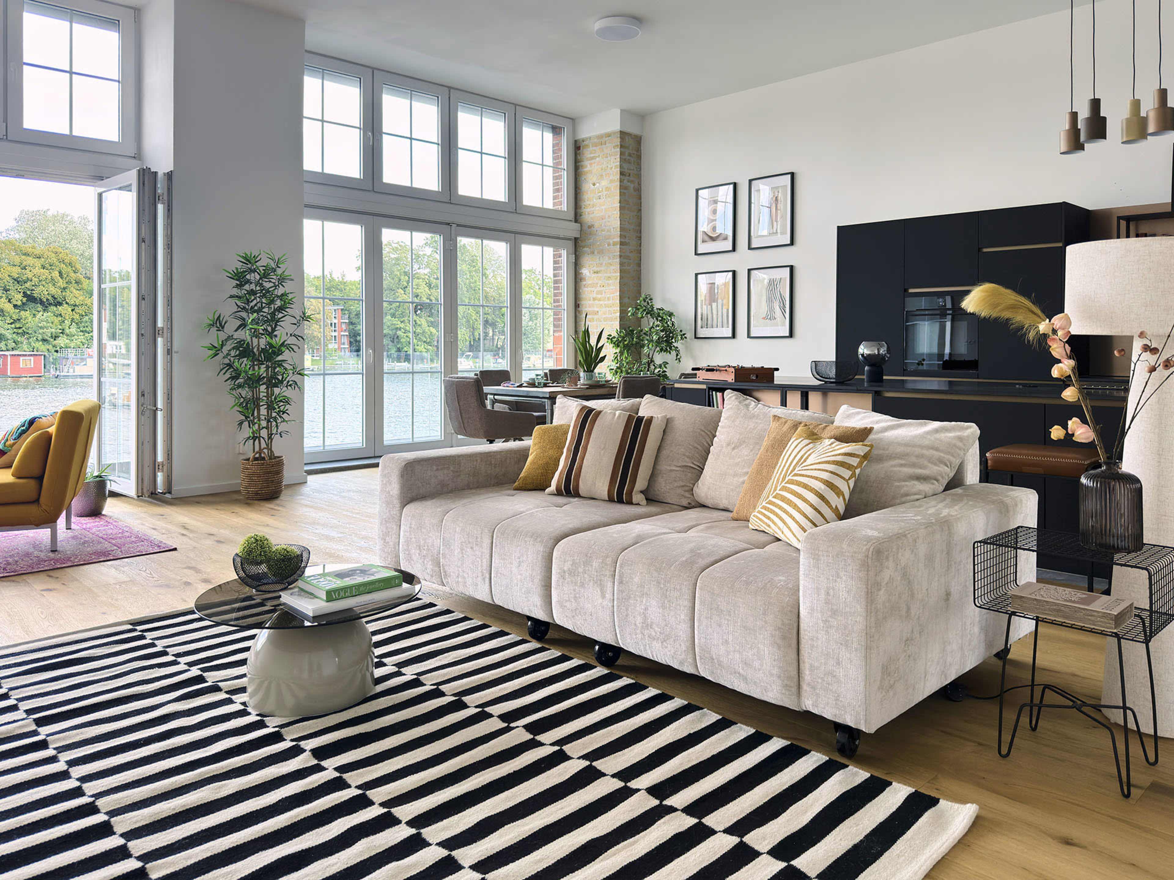 The image shows a modern living room with a light-colored sofa, a striped area rug, and large windows that offer a view of the water.