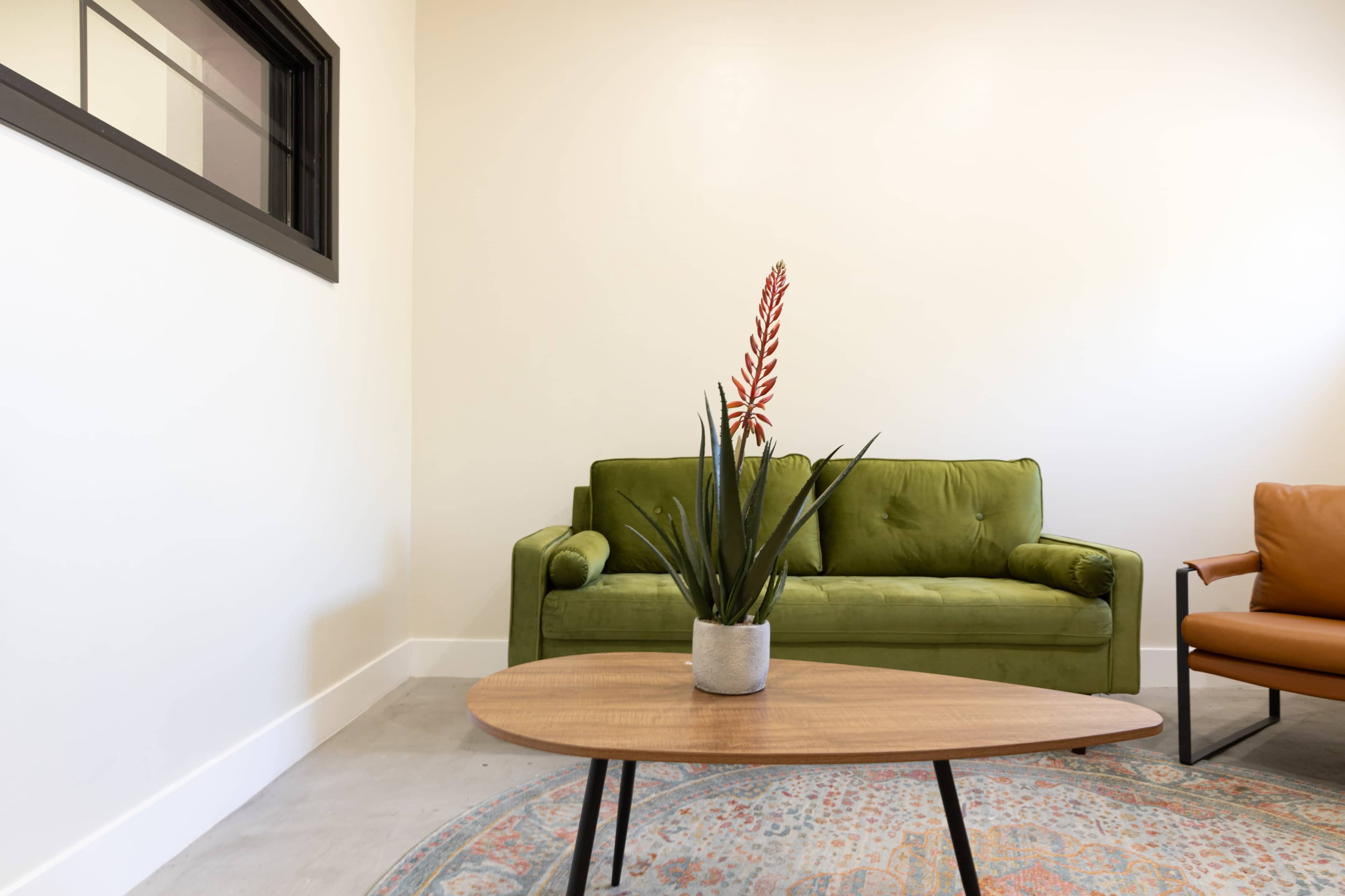A green couch and an orange chair arranged around a wooden coffee table with a potted plant on it, set in a modern room with light-colored walls.