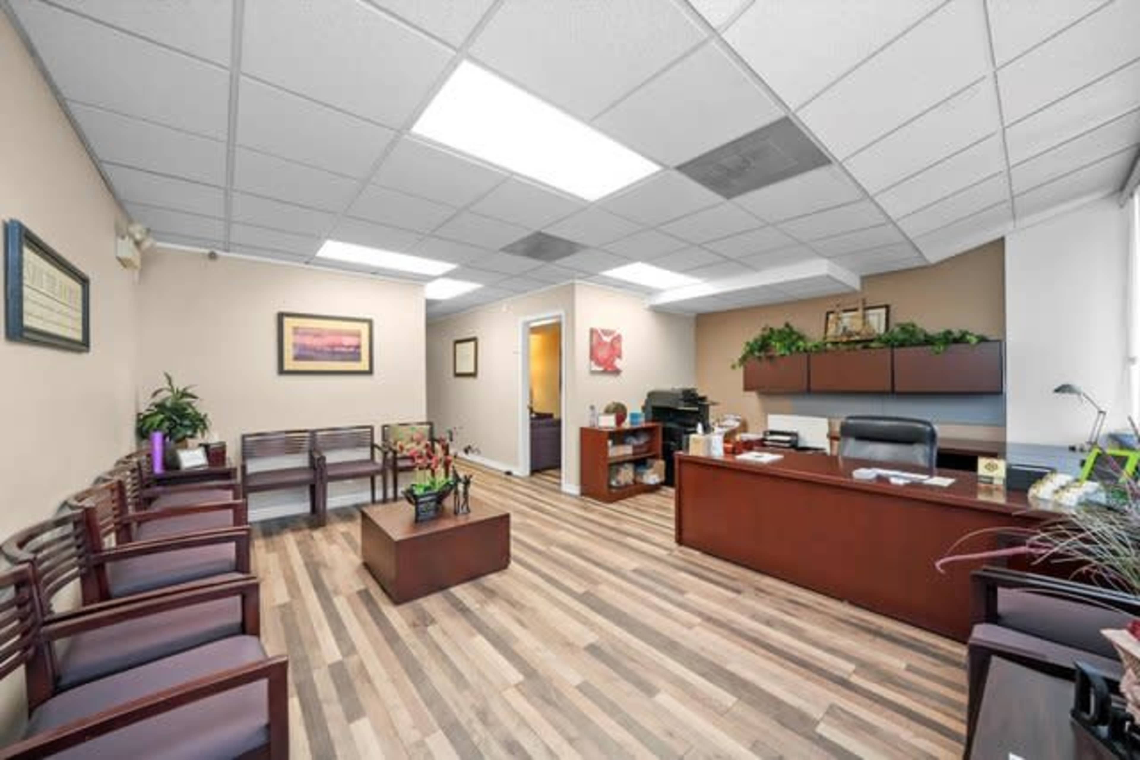 The image shows a modern office reception area with a wooden desk, several chairs along the wall, and a plant arrangement in the center.