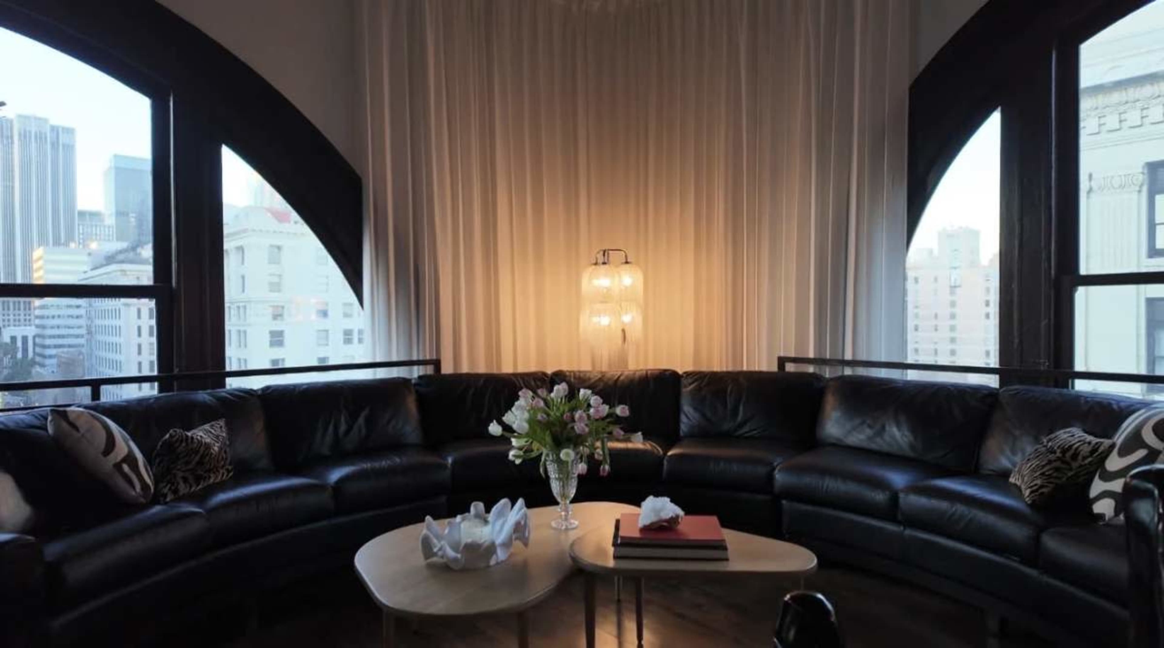 A curved leather sectional sofa surrounds a coffee table with a floral arrangement in a well-lit room featuring large arched windows.