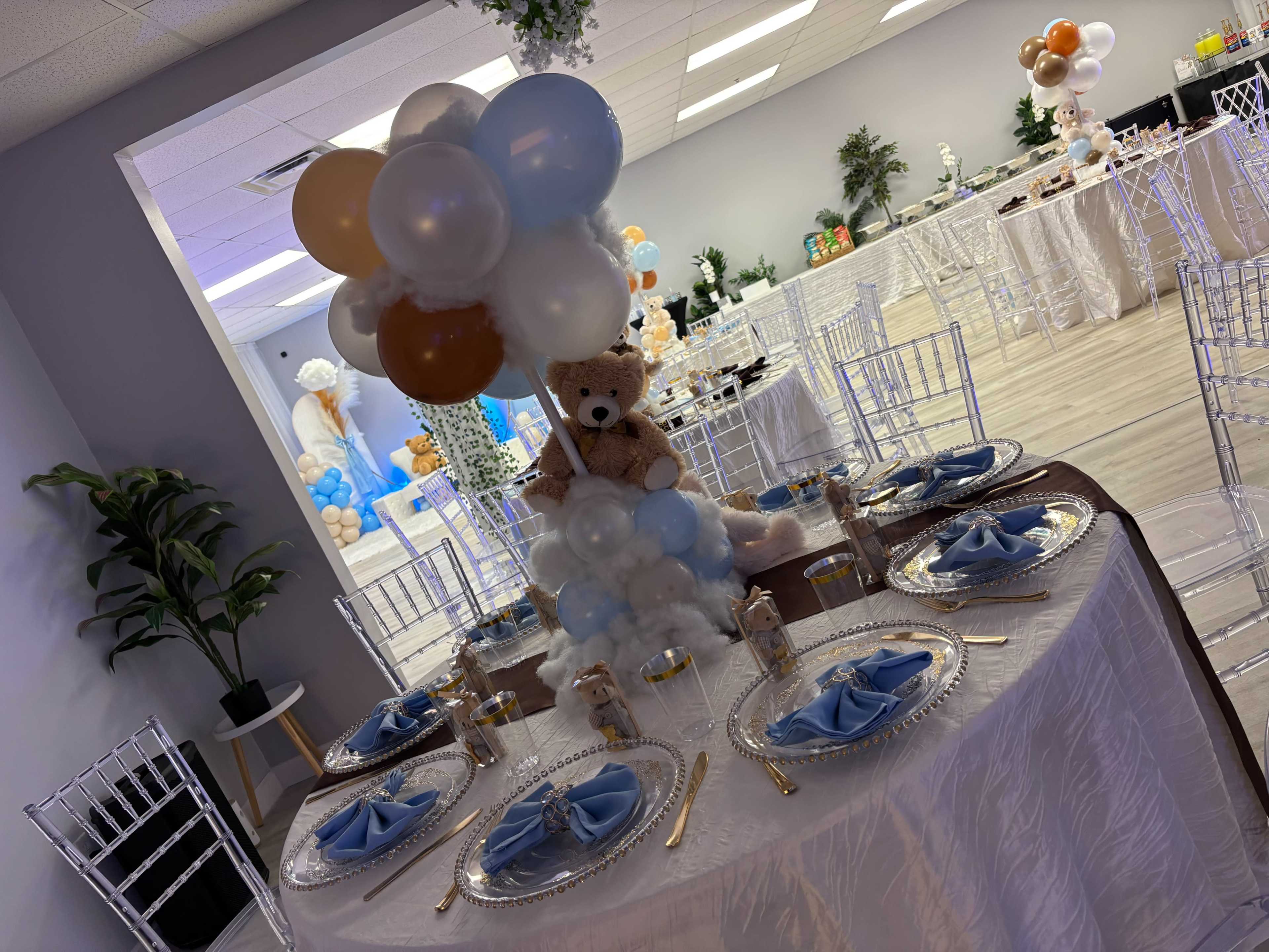 A decorated table is set with blue napkins and clear plates, featuring a teddy bear centerpiece amidst balloons, in a spacious event room with additional tables in the background.