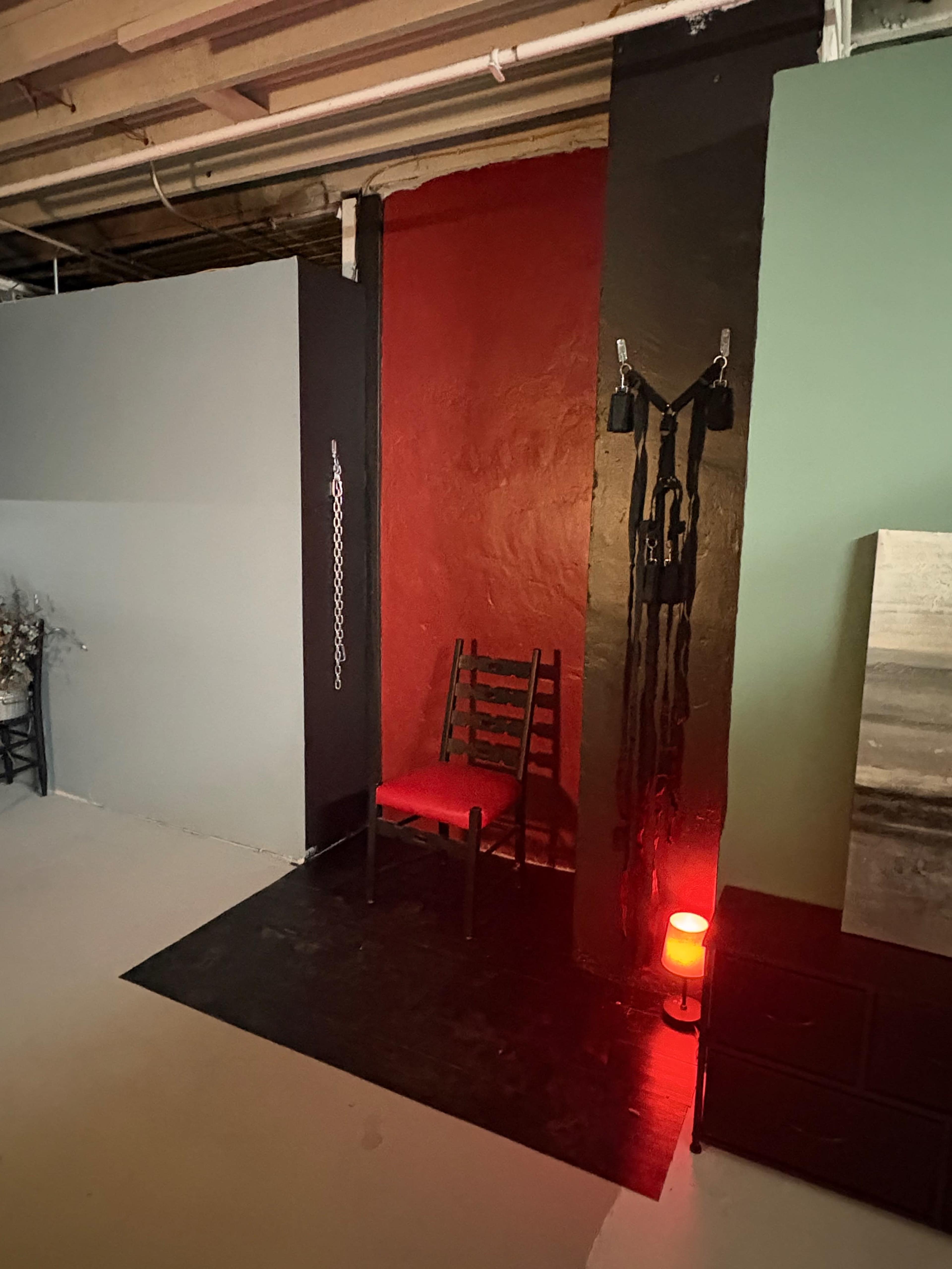 The image shows a dimly lit room featuring a red wall with a black accent, a wooden chair, and various accessories hanging nearby, including restraints, next to a low red light.