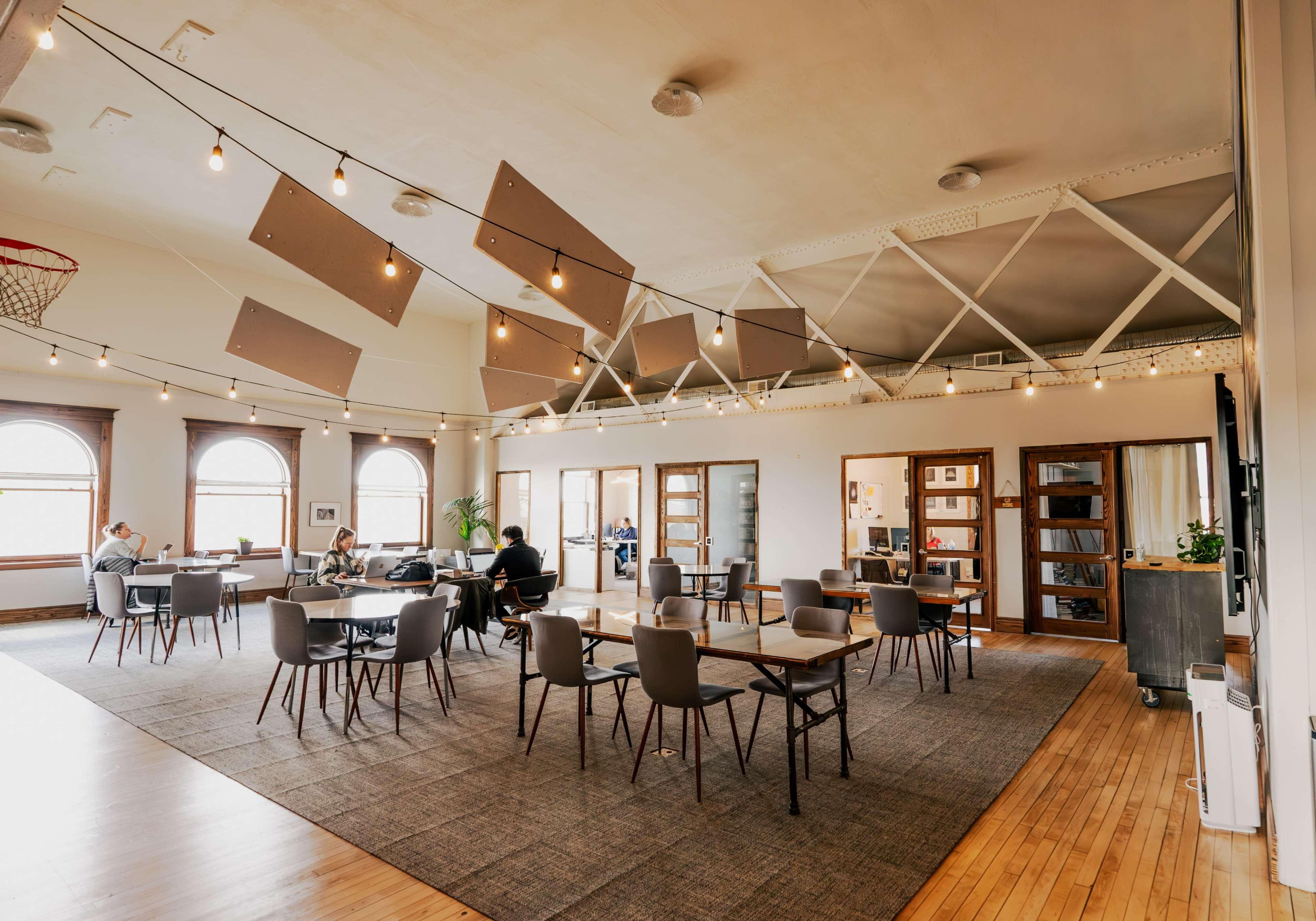A spacious room with tables and chairs arranged for dining or working, featuring large windows and string lights overhead.