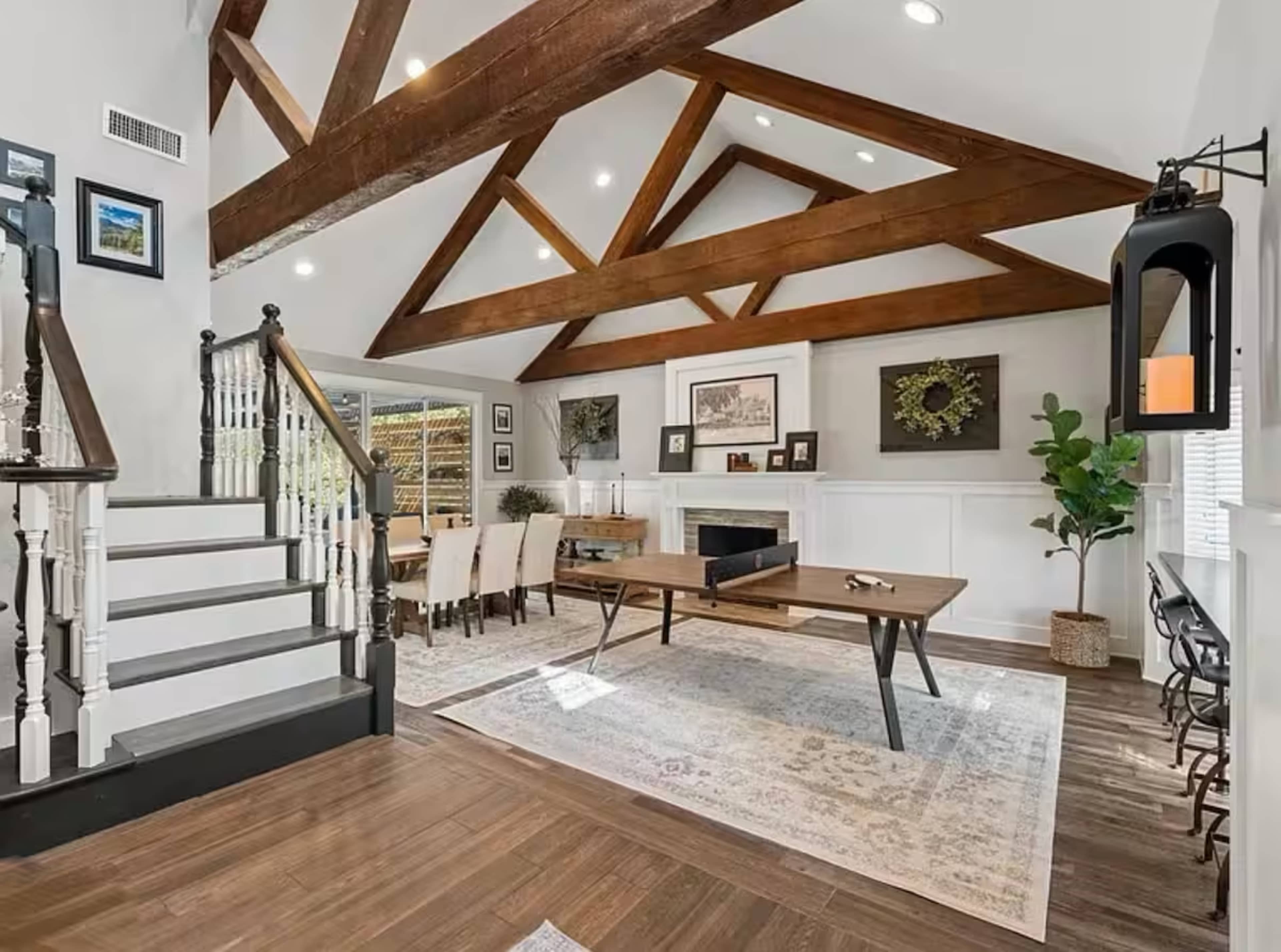 The image features a spacious living area with a high beamed ceiling, a central table, and a staircase leading to an upper level, accented by decorative walls and a large window.