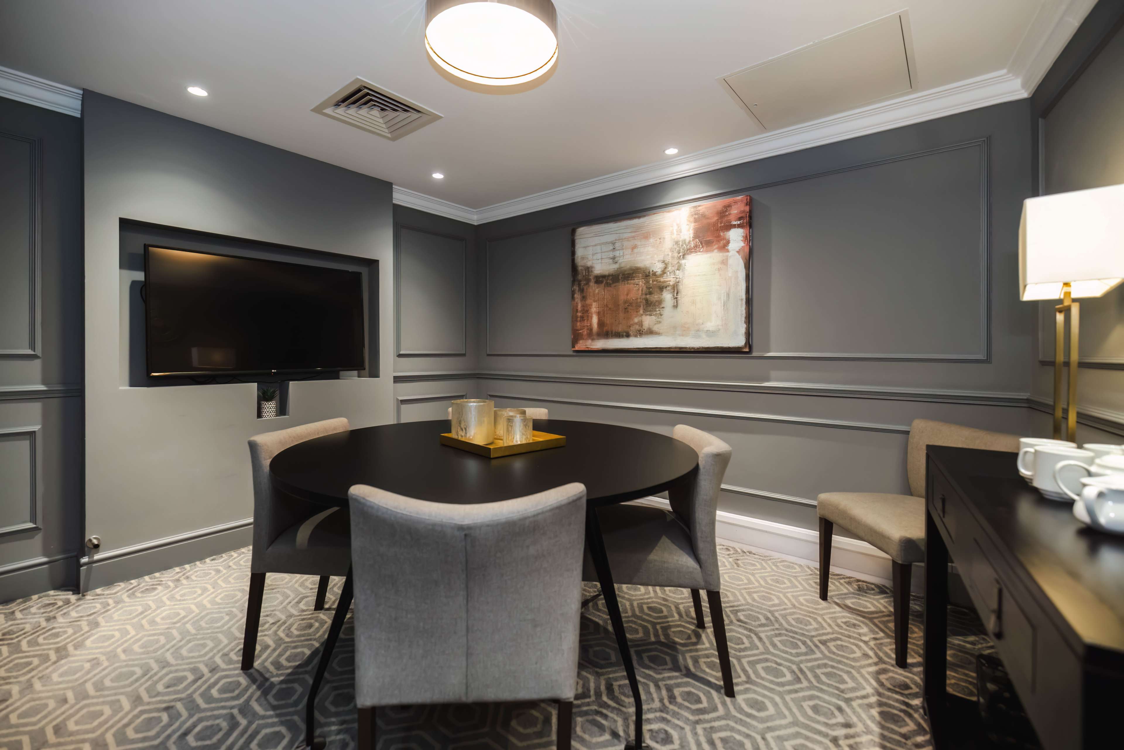 A small dining area with a round black table surrounded by four grey chairs, a wall-mounted television, and an abstract painting on the wall.