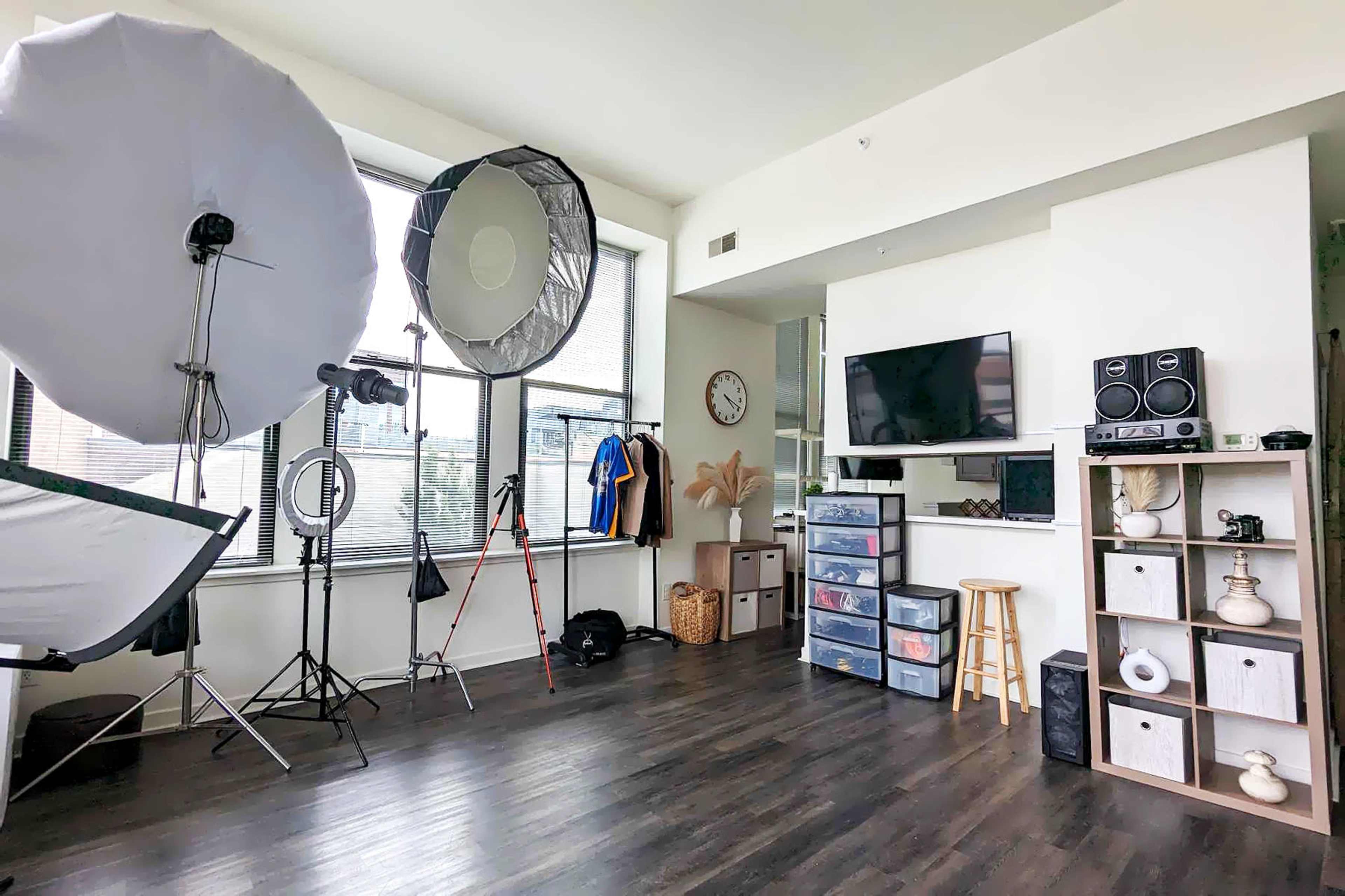 A well-lit studio space with various photography equipment, including large softboxes, a tripod, and storage units, along with a television mounted on the wall.