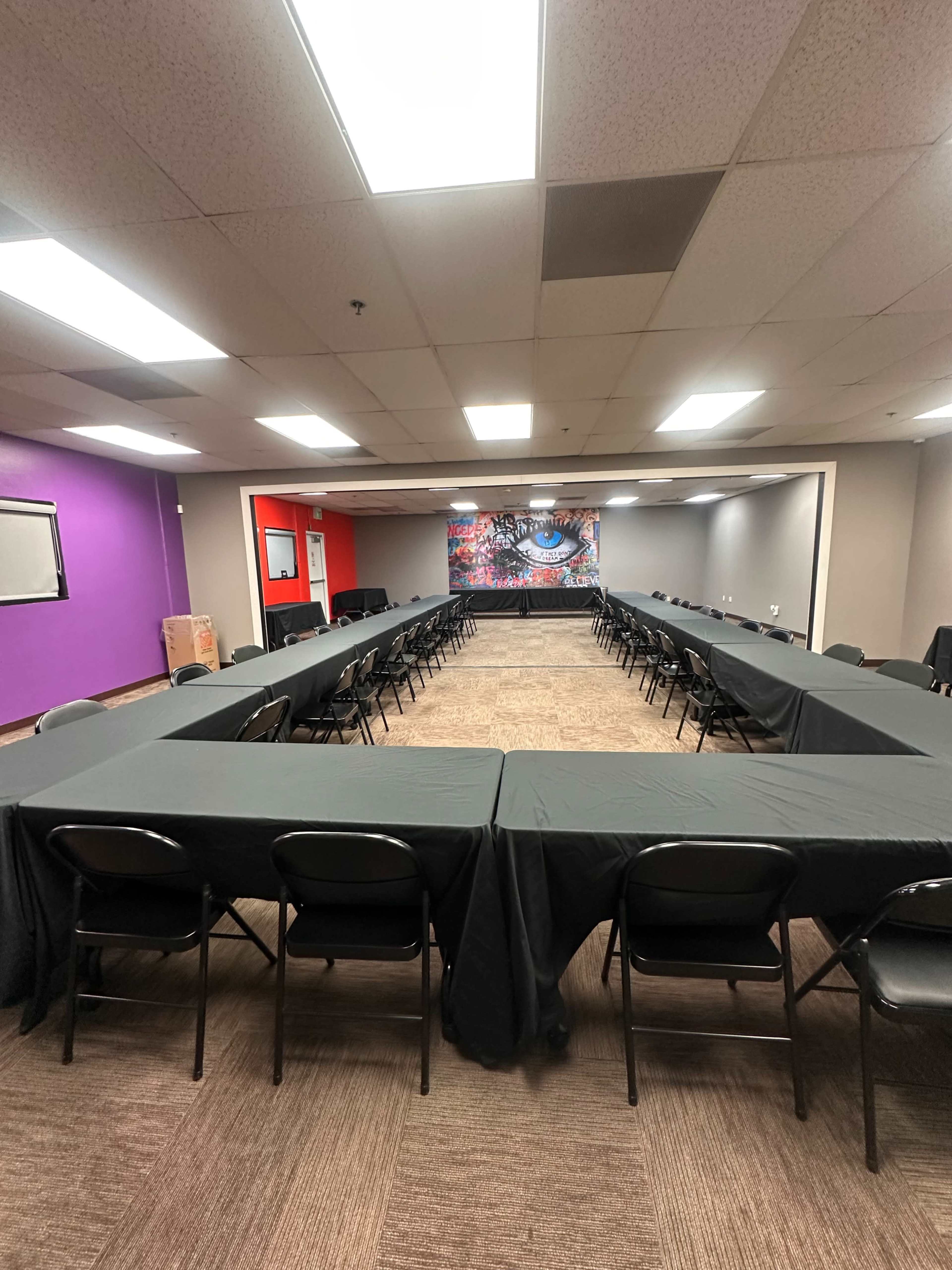 The image shows a room arranged with multiple tables in a U-shape, surrounded by chairs, and featuring a colorful mural on one wall.