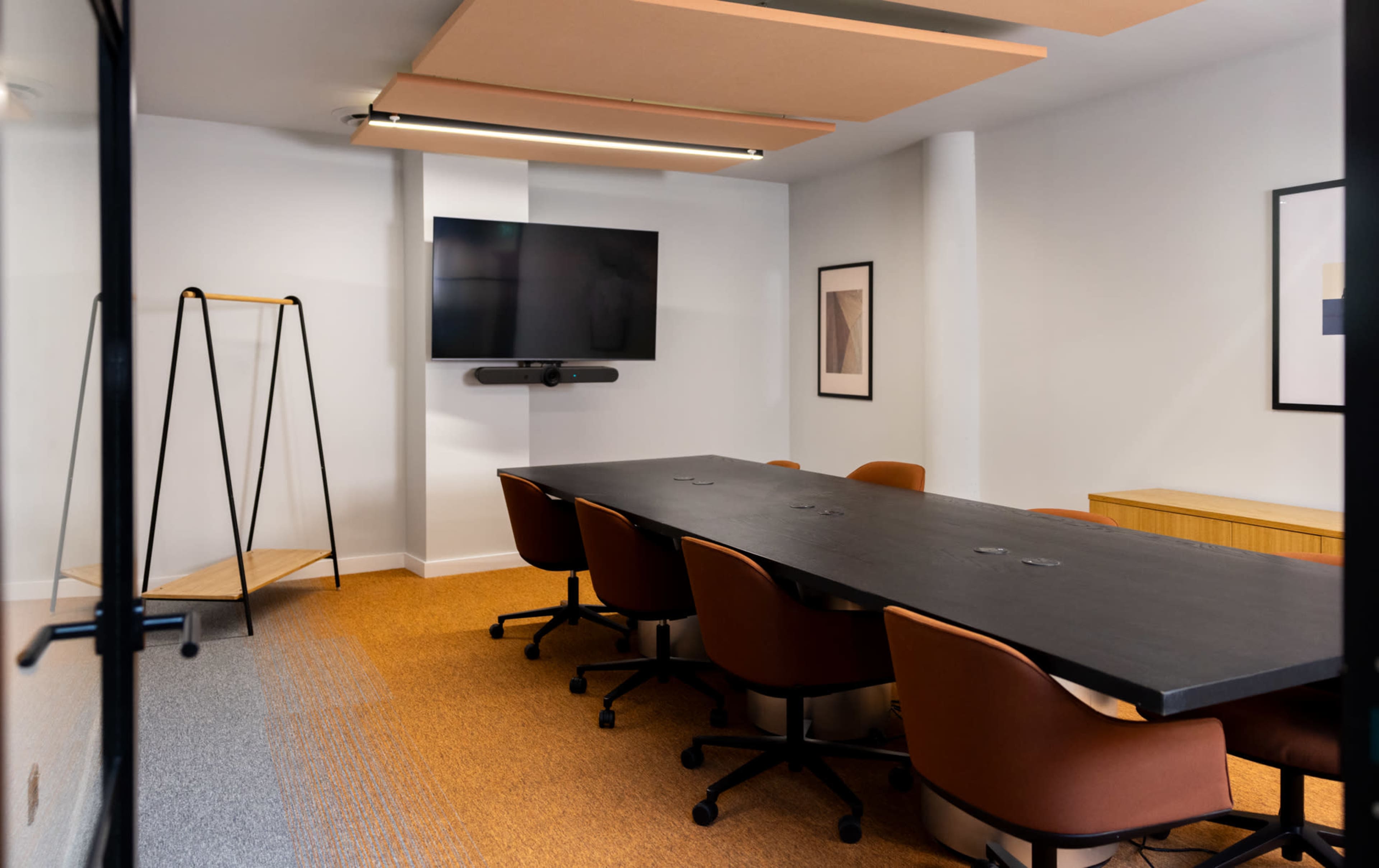 A modern conference room features a long black table surrounded by brown swivel chairs and a mounted television on the wall.