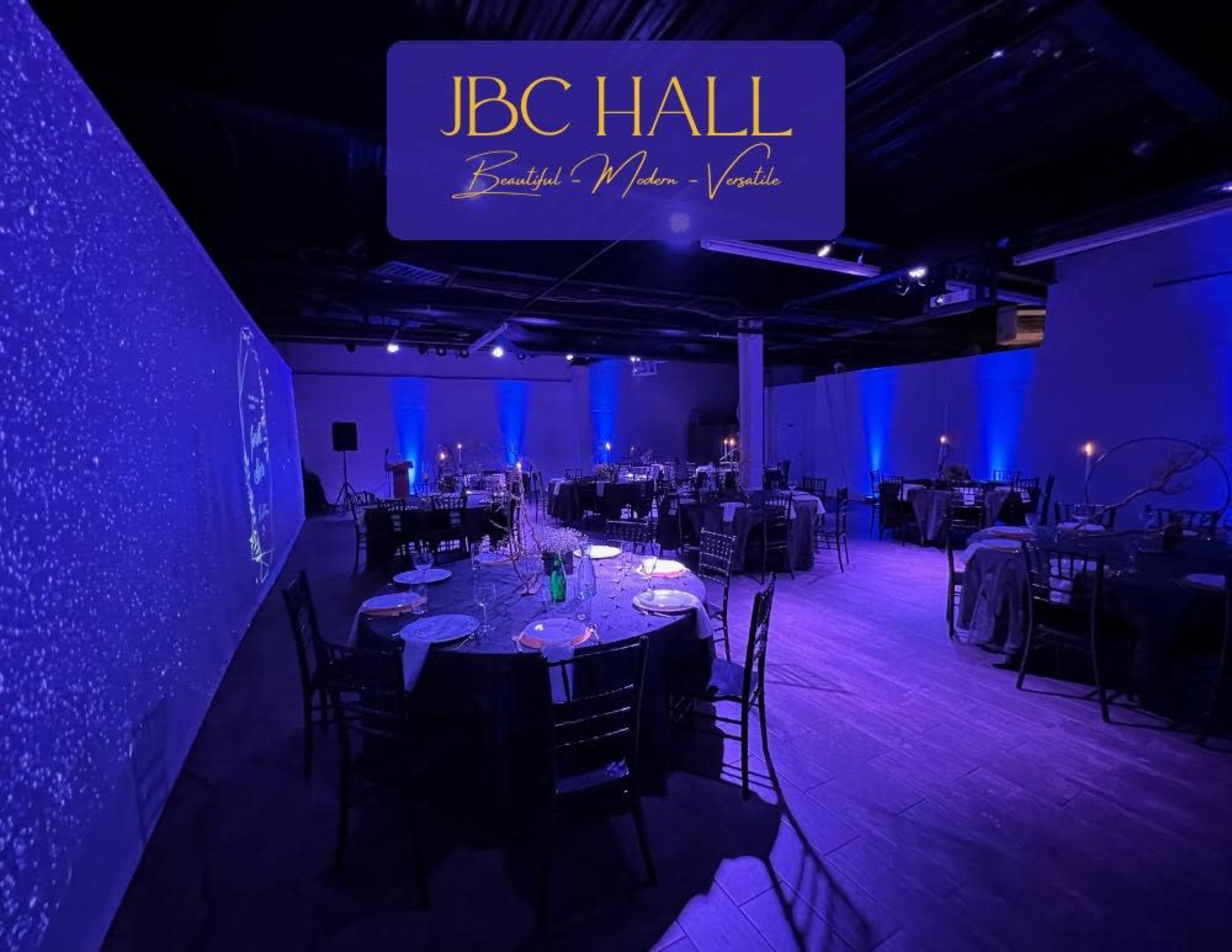 The image shows a spacious event hall set up for a formal gathering, featuring elegantly arranged tables with dark chairs and ambient blue lighting.