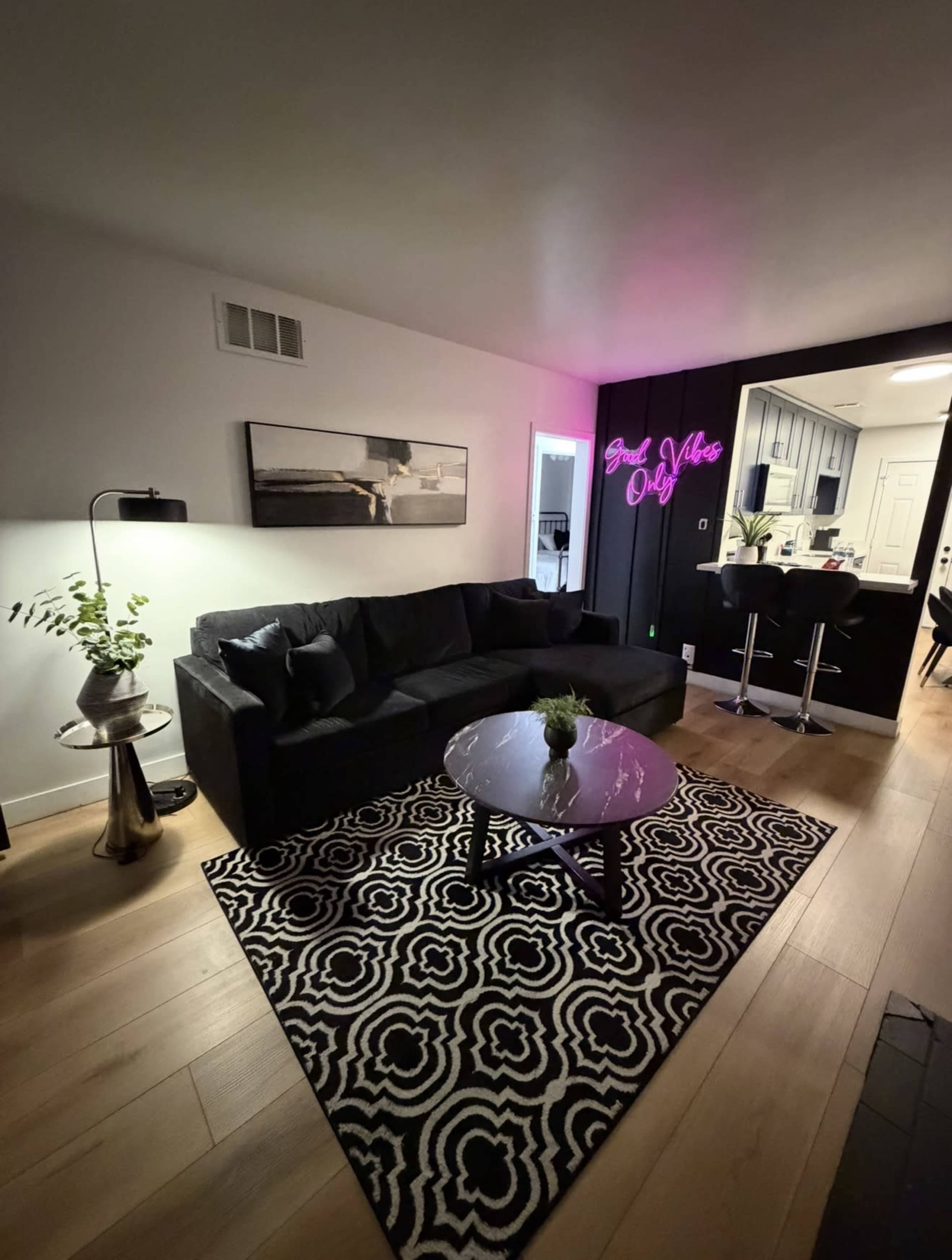The living room features a black sofa and a round glass coffee table, with a patterned rug underneath and a neon sign on the wall.