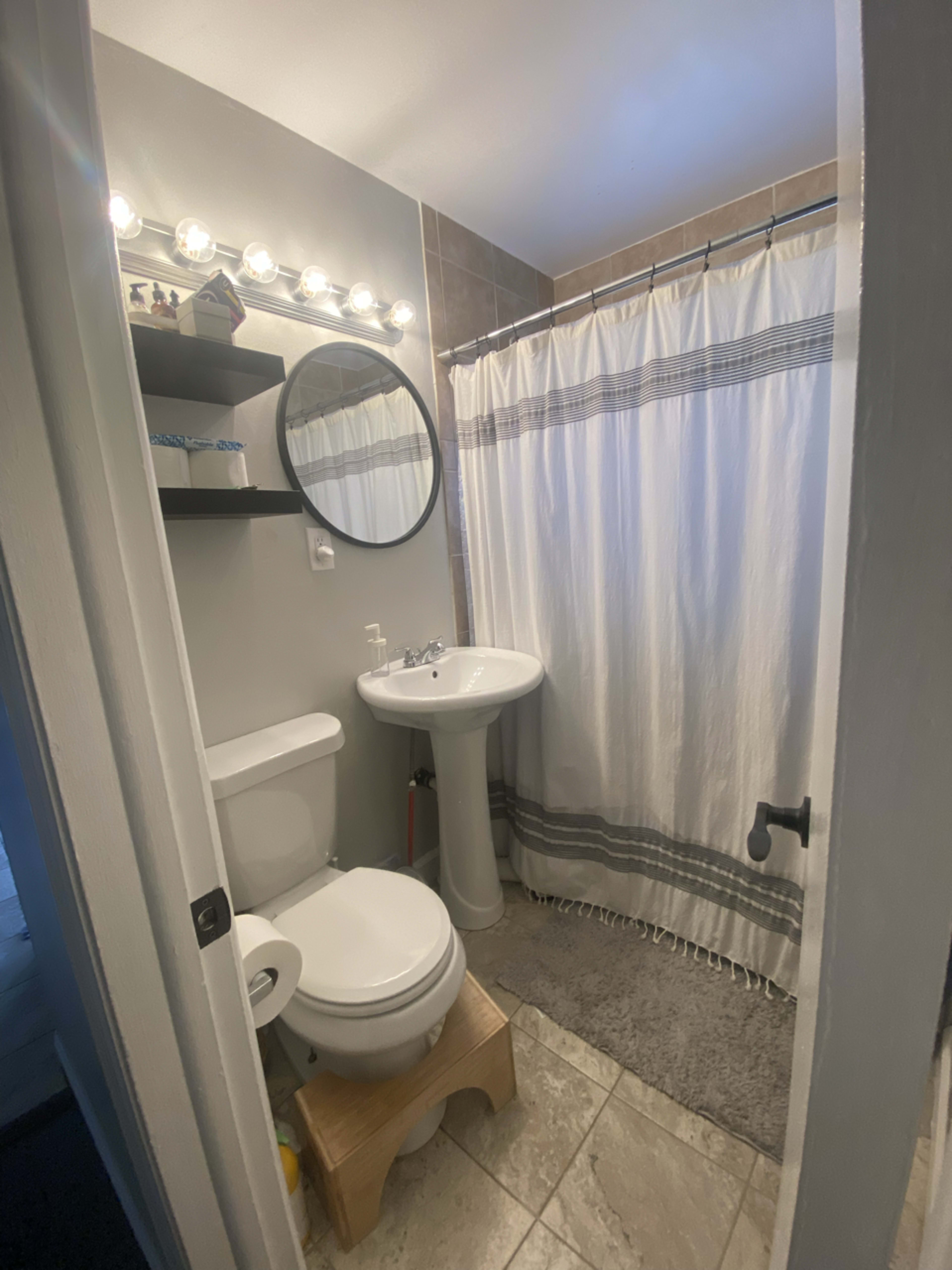A small bathroom features a shower with a white curtain, a pedestal sink, a toilet, and a wooden stool on tiled flooring.