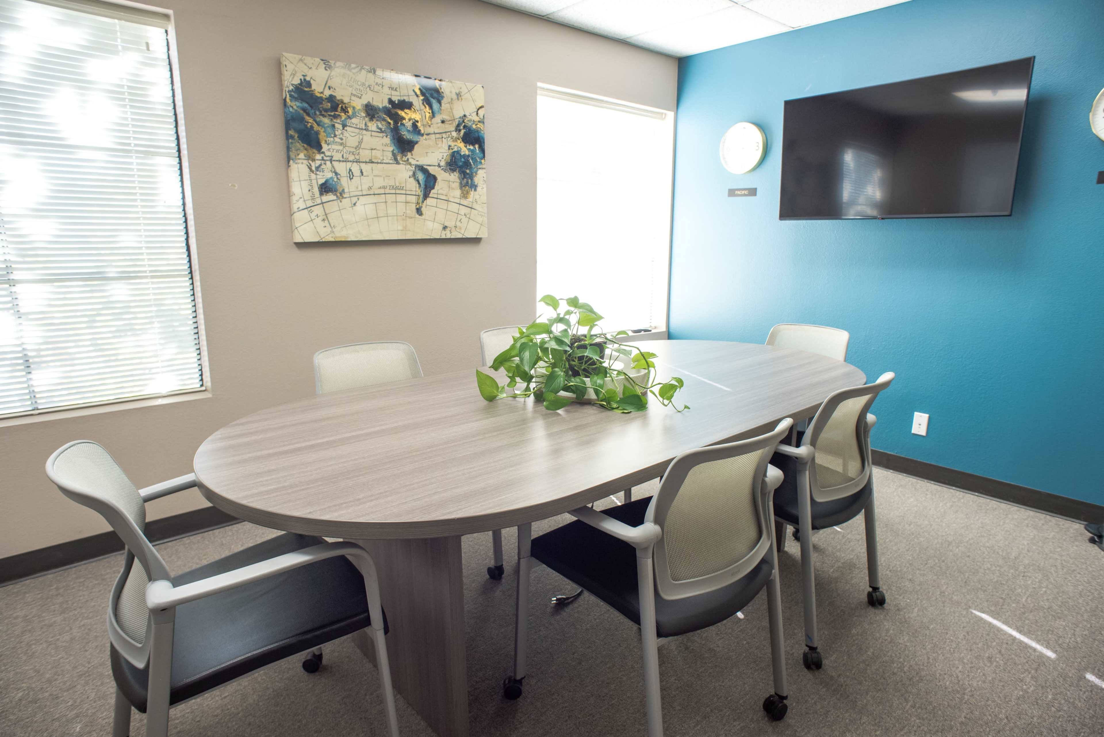 A modern conference room features an oval table surrounded by six chairs, with a large wall-mounted television and a world map artwork.