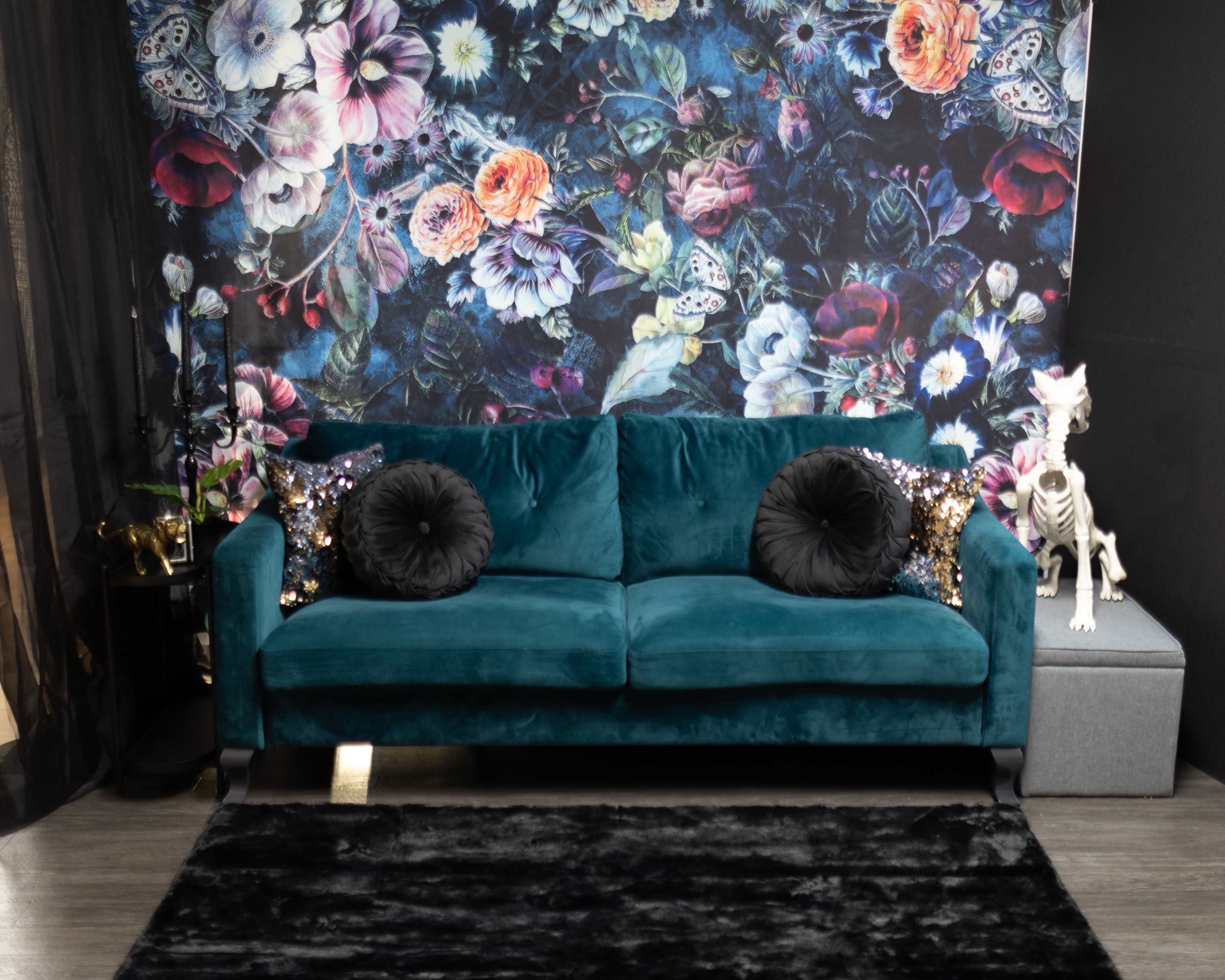 A teal sofa with black decorative pillows sits in front of a floral wallpaper and is accompanied by a decorative skeleton and a dark rug.