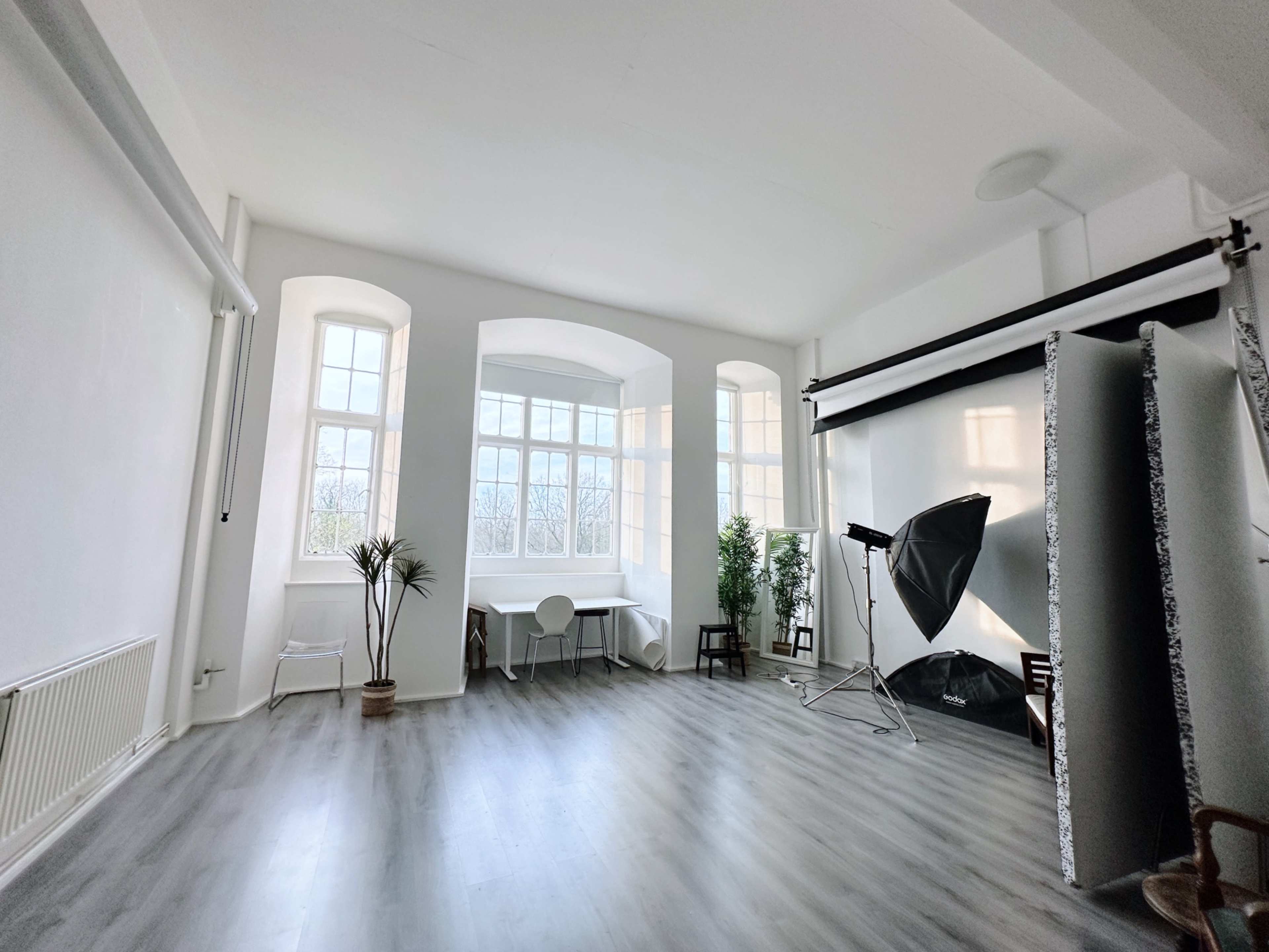 The image shows a bright, empty studio space with large windows, light wood flooring, and photography equipment set up in one corner.