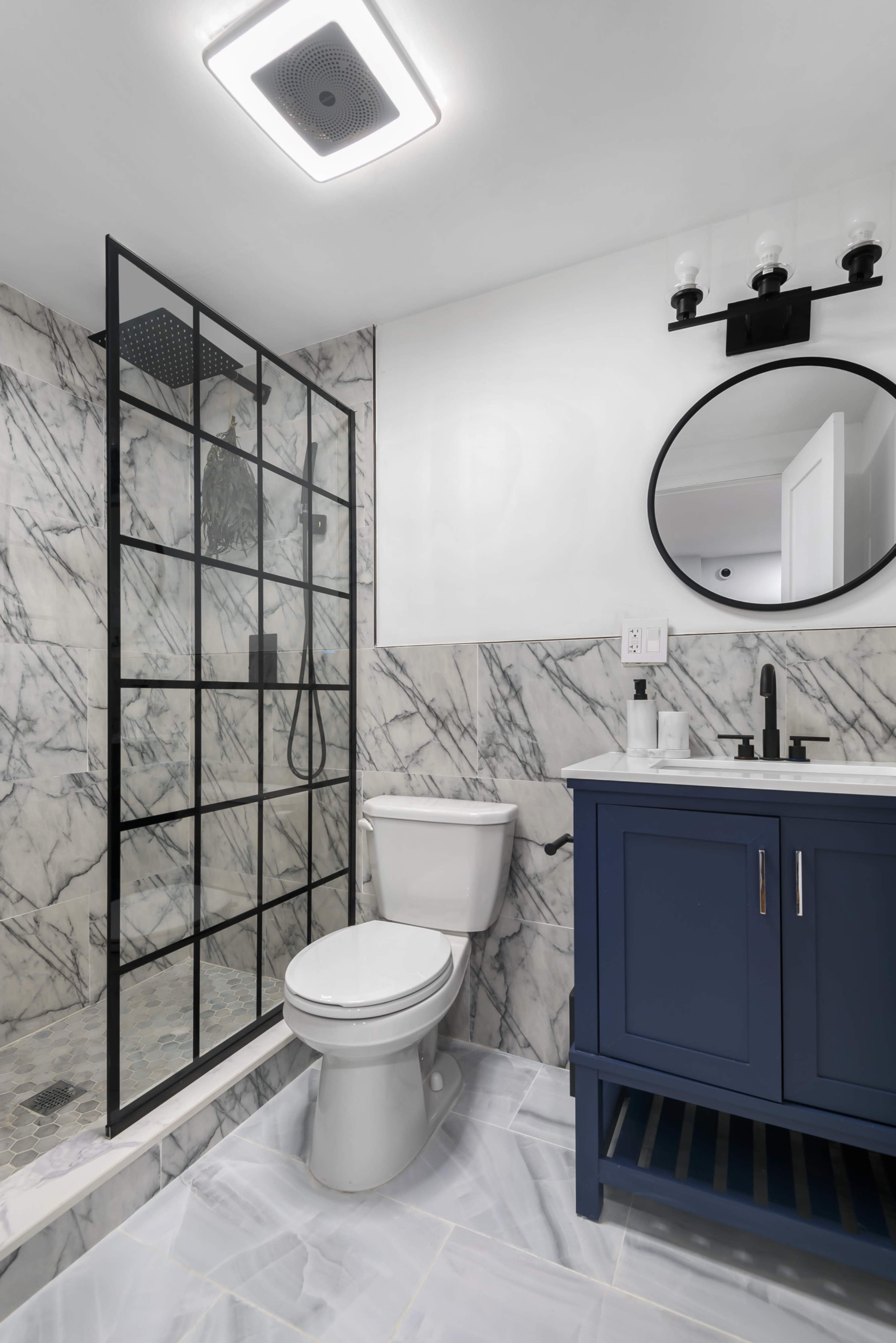 A modern bathroom featuring a glass shower enclosure, a toilet, and a blue vanity with a round mirror above it, set against marble walls.