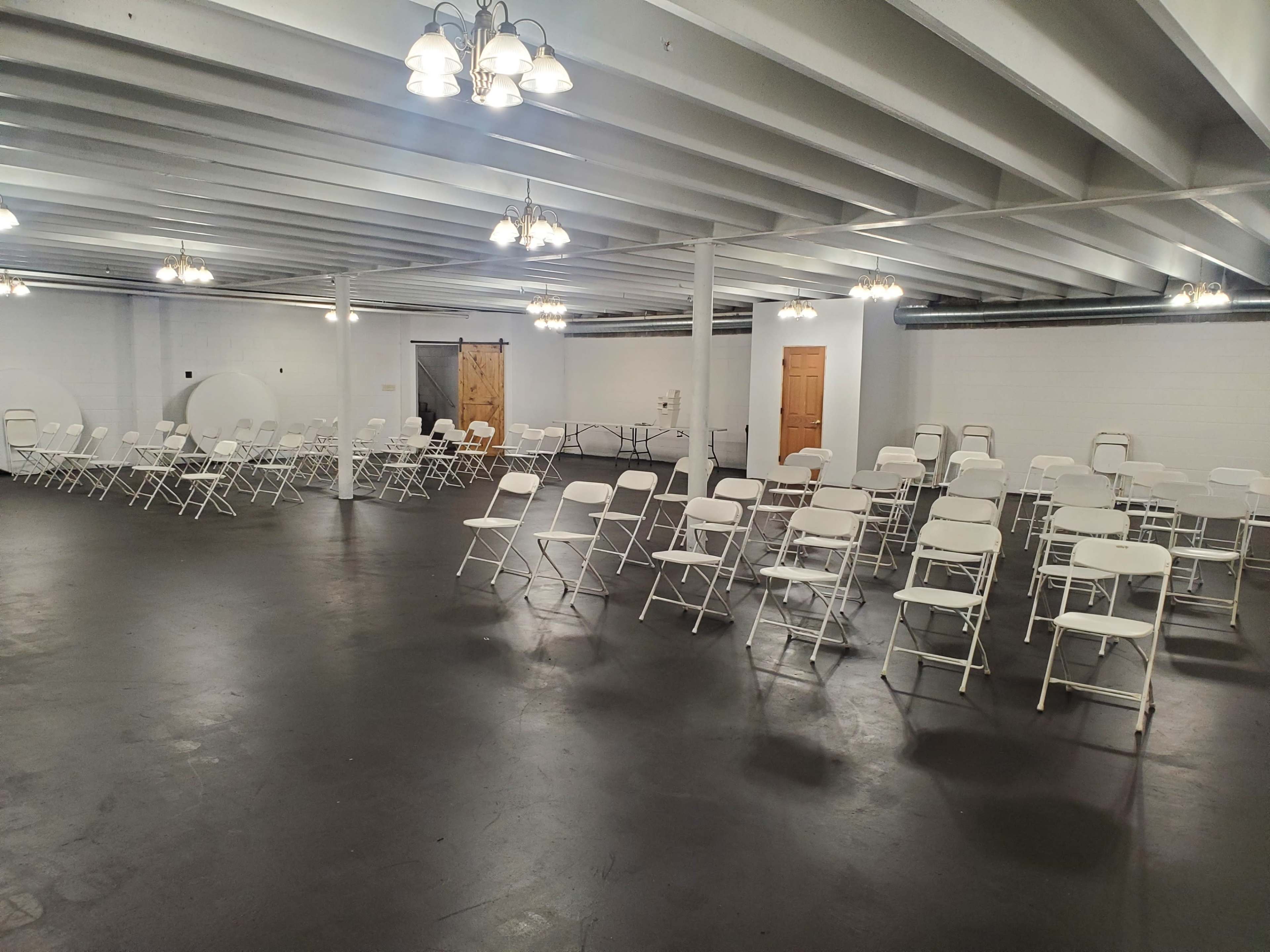 A large, empty room arranged with rows of white folding chairs under overhead lights.