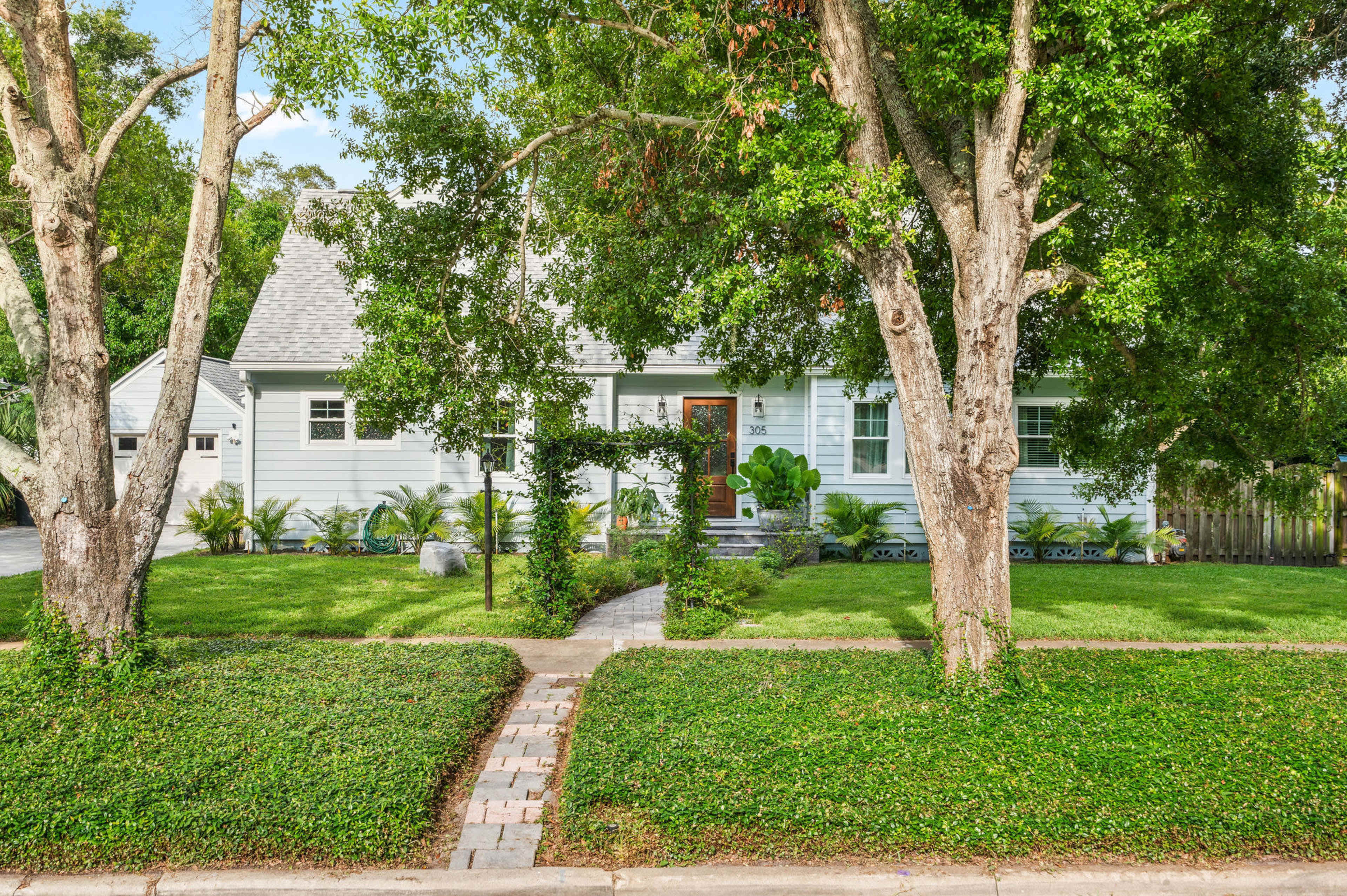 The image shows a light blue house with a steep roof, surrounded by lush green landscaping and framed by two large trees.