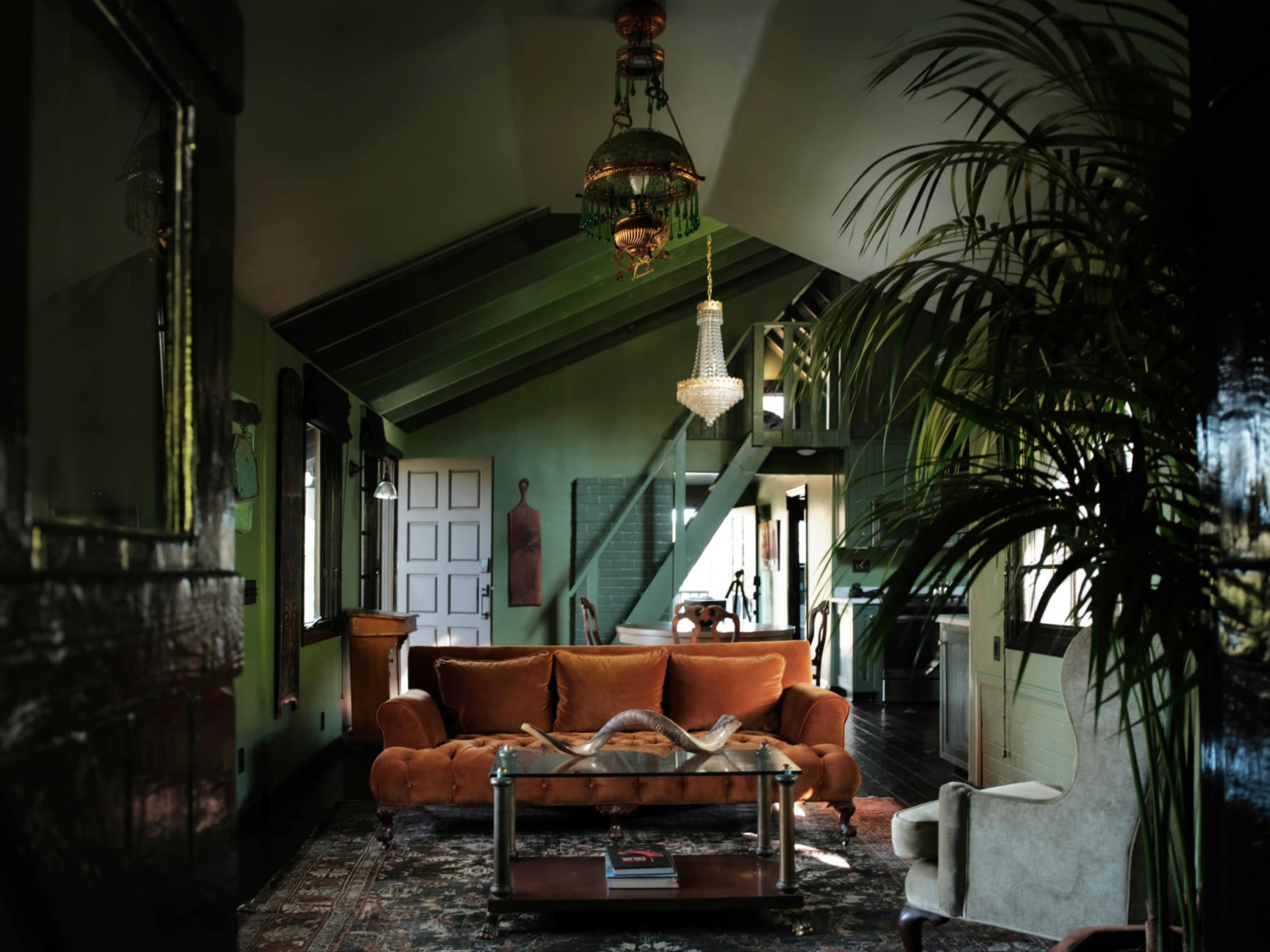 The image depicts a spacious living room featuring a large orange couch, a glass coffee table, and decorative plants against a green-painted wall.
