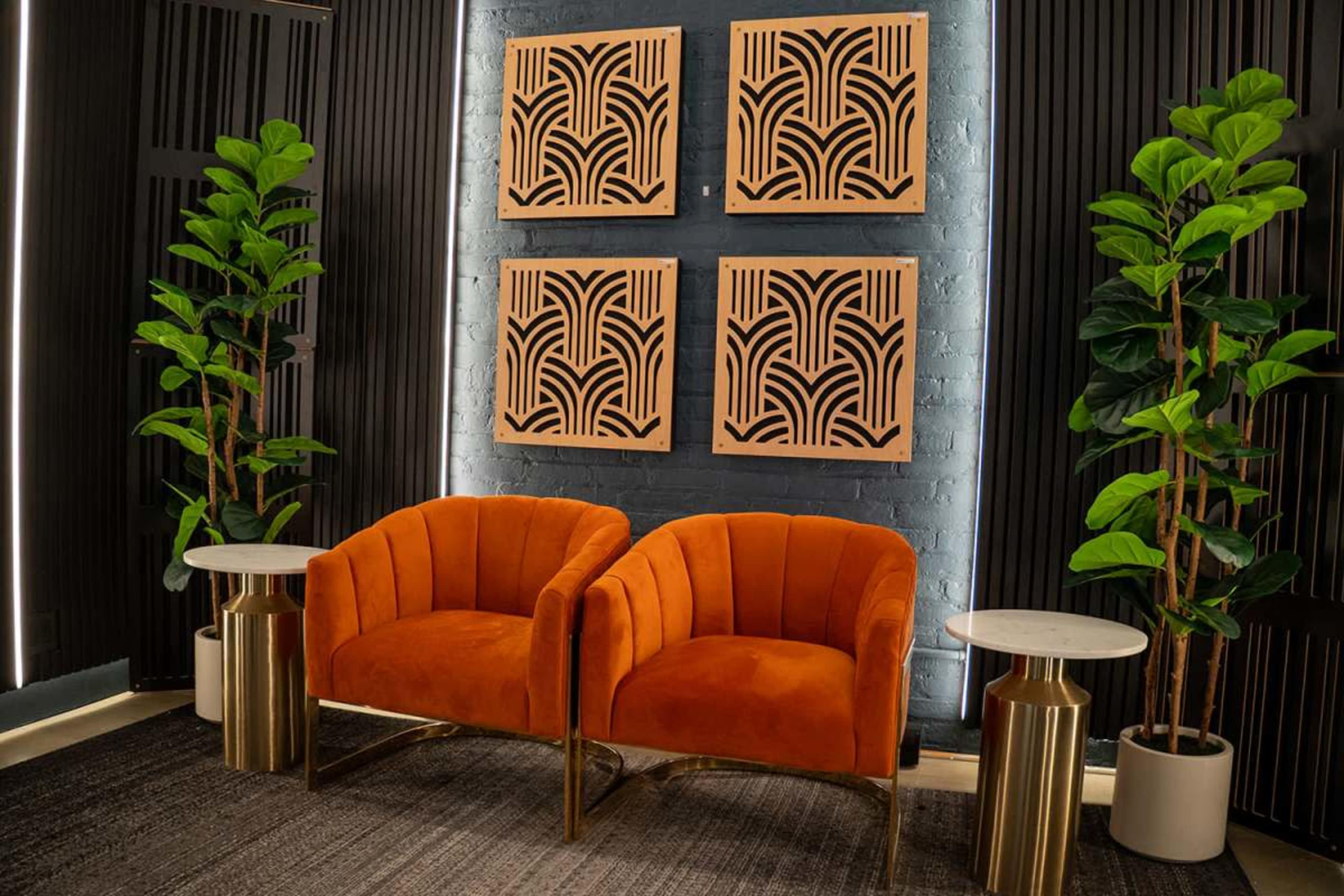 The image features two orange upholstered chairs paired with white marble-topped side tables, set against a backdrop of abstract wall art and leafy green plants.