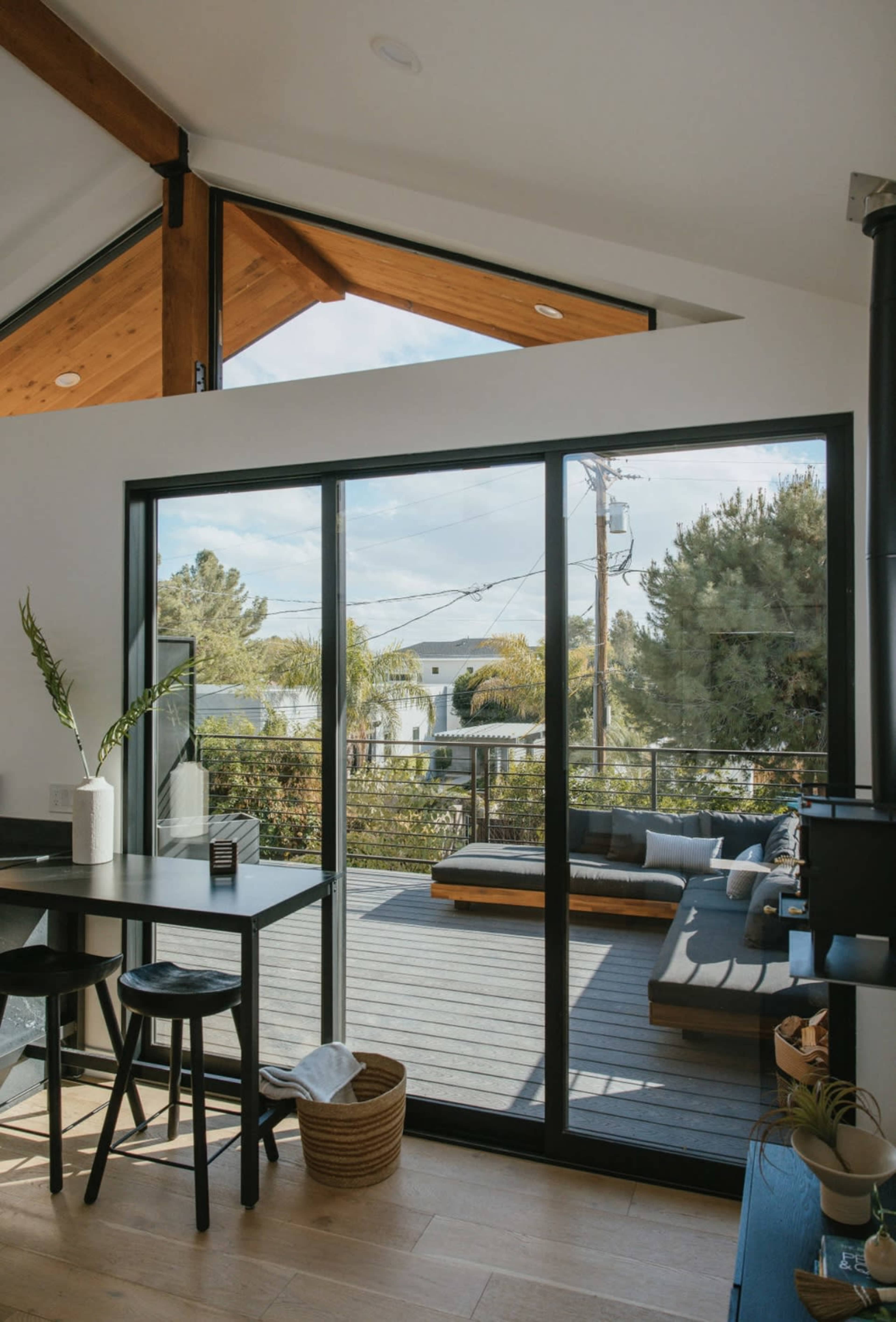 A modern interior space features large glass doors opening to a wooden deck with outdoor seating, framed by greenery and a clear sky.