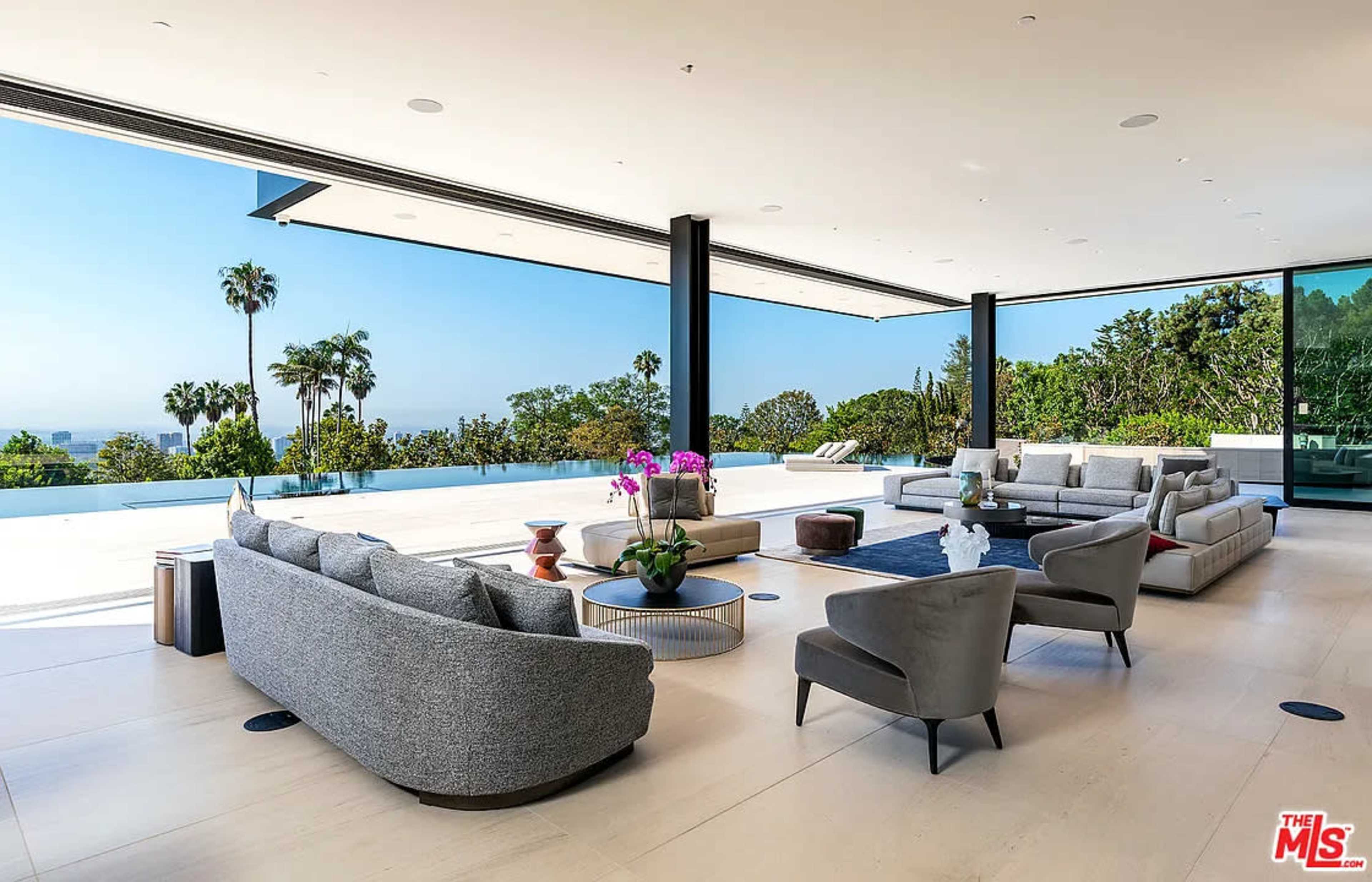 The image shows a modern living area with large glass walls offering a view of green trees and a pool, furnished with gray sofas and a coffee table.