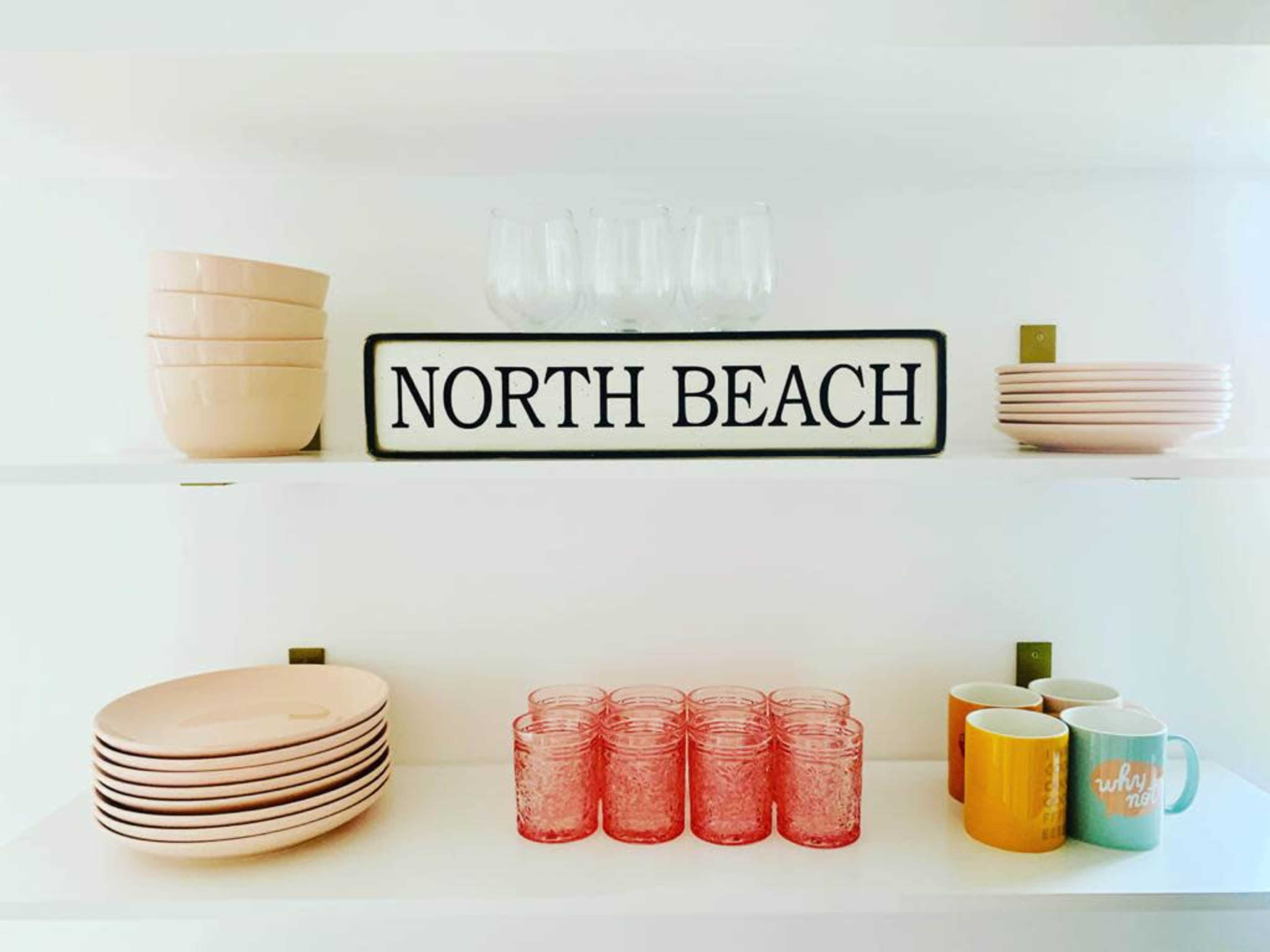 A white shelf displaying various kitchenware, including bowls, plates, cups, and glasses, along with a sign reading "NORTH BEACH."