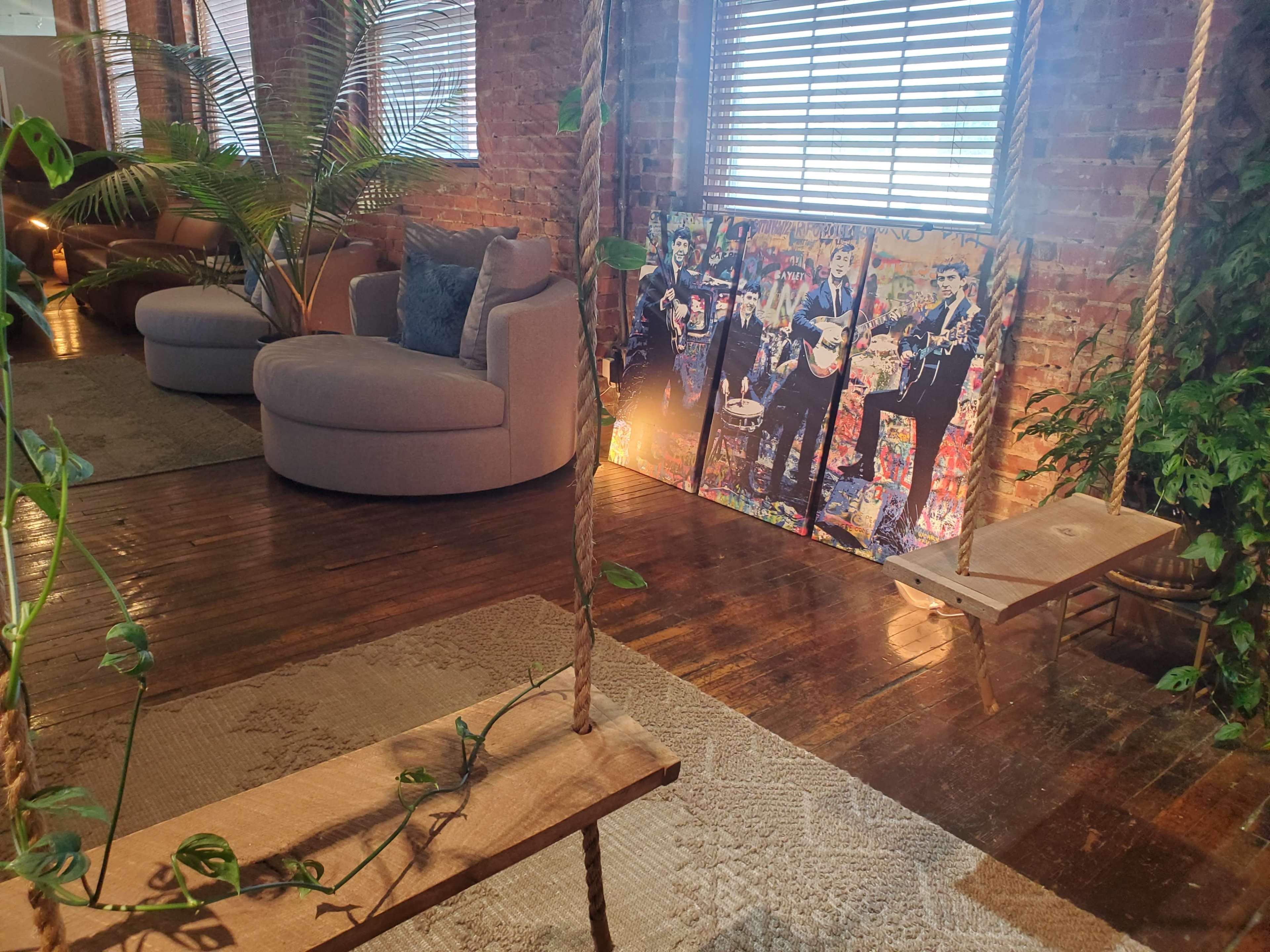 The image shows a room with a round gray sofa, a wooden swing, a large colorful painting of musicians, and plants positioned near brick walls and windows.