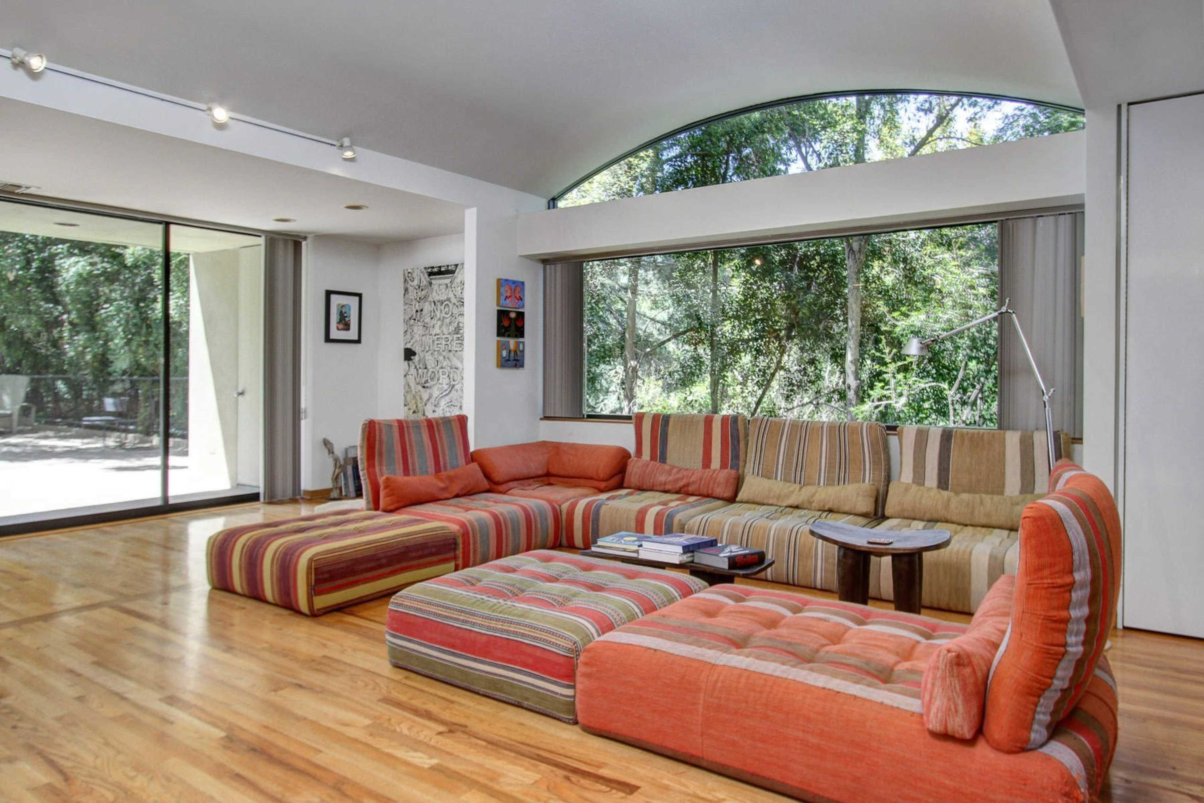 A spacious living room features a large, striped sectional sofa, wooden floors, and large windows overlooking greenery.