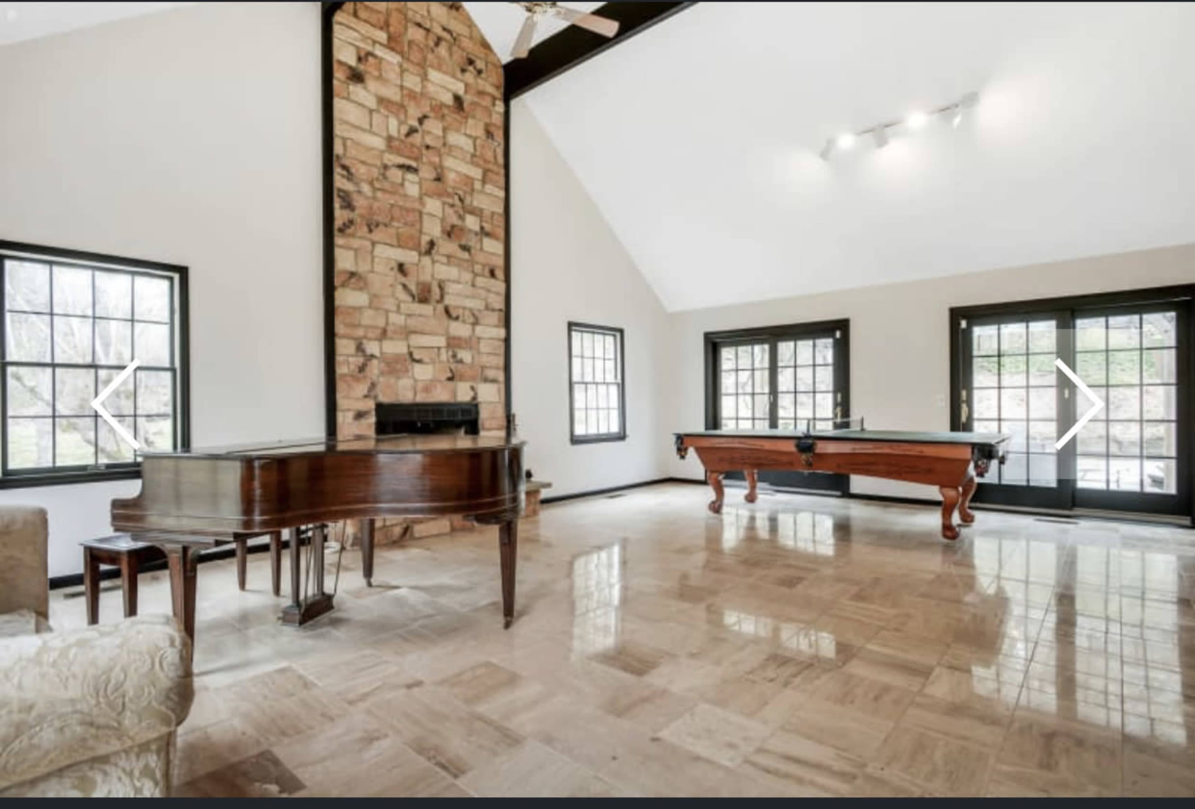 The room features a stone fireplace, a piano, and a billiard table, with large windows allowing natural light to enter.