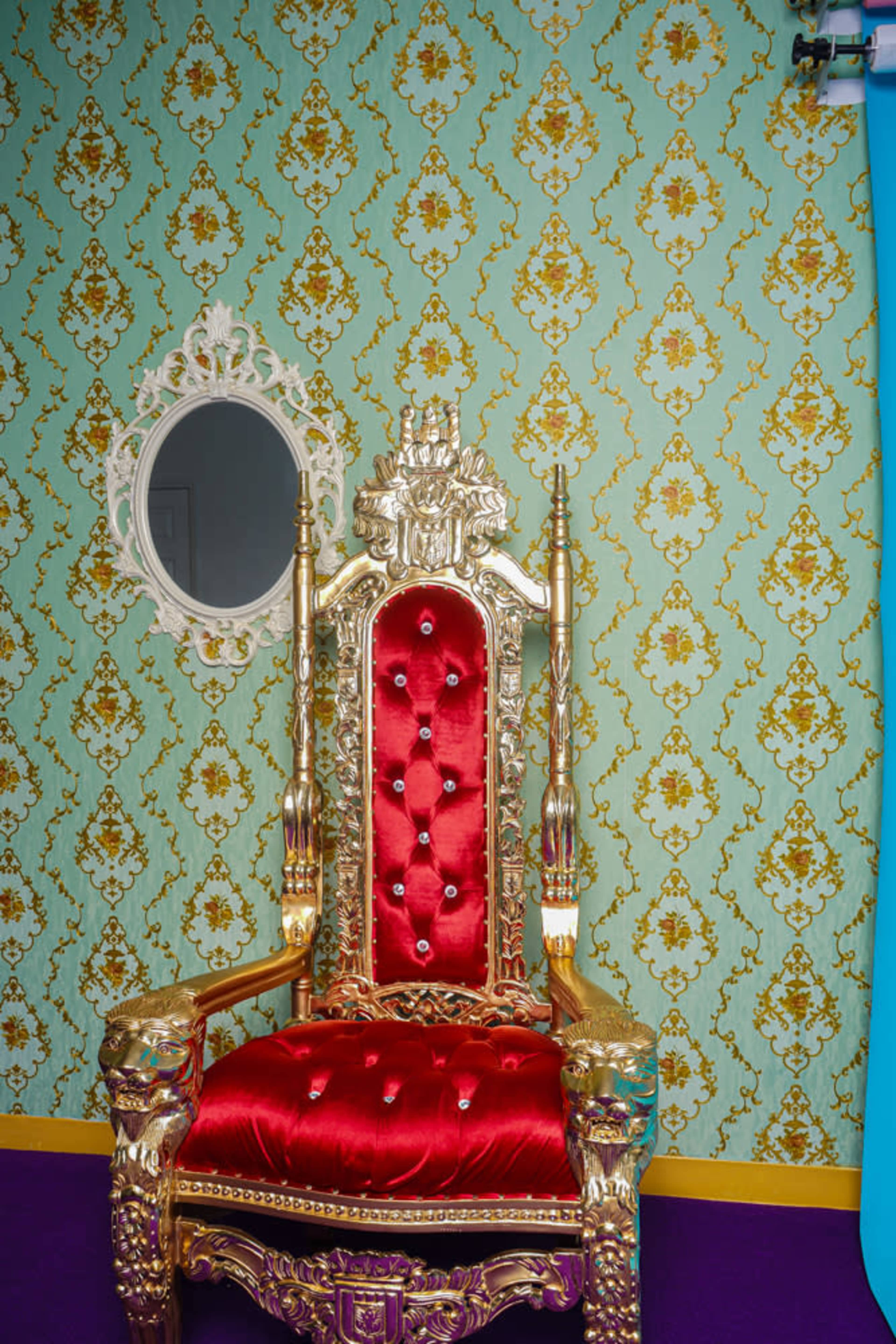 A gold and red throne is positioned against a patterned green wallpaper, with a circular mirror hanging on the wall nearby.