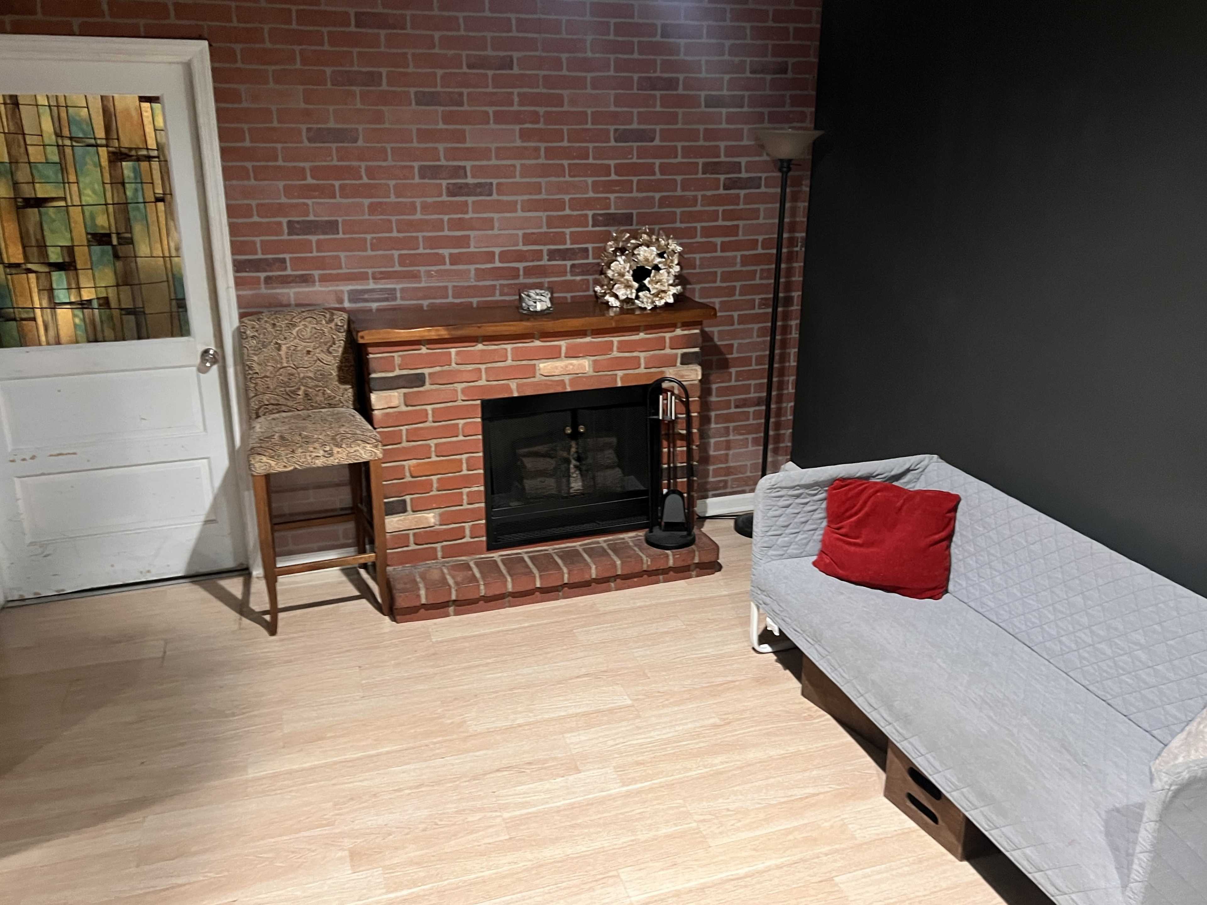 The image shows a cozy room with a brick fireplace, a gray couch covered with a blanket, and a bar stool near a door with stained glass.